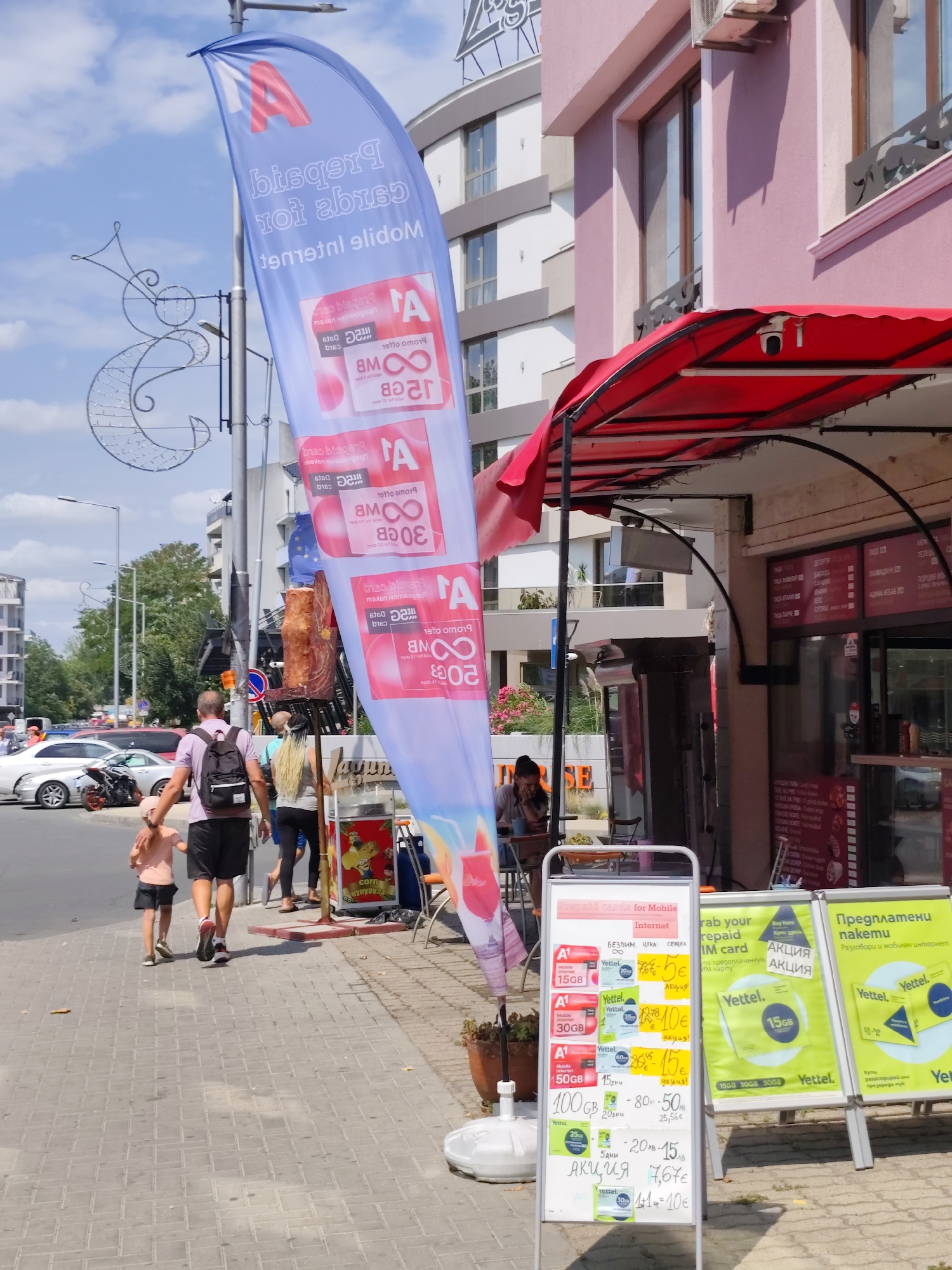 The image shows a street scene in Nesebar, Bulgaria. In the foreground, a tall, blue flag with white lettering and a red "A" logo is prominent, advertising "Data Internet cards for Mobile". To the right of the flag, a white A-frame sign displays offers for mobile internet packages, including different data amounts (15GB, 30GB, 50GB, 100GB) and prices in Euros and Leva, with "Акція" (Action/Offer) highlighted. Further to the right, another green A-frame sign advertises "Yettel" mobile plans.
A group of people is walking on the sidewalk. A man in a purple shirt and black shorts, wearing a backpack, is holding the hand of a young child in a pink shirt and shorts. Behind them, a woman with long blonde hair is walking.
In the background, a pink building with windows and a red awning is visible, possibly a shop or restaurant. The street has cars parked and driving. The environment appears to be a commercial area on a sunny day. The overall impression is of a street with businesses advertising mobile services.