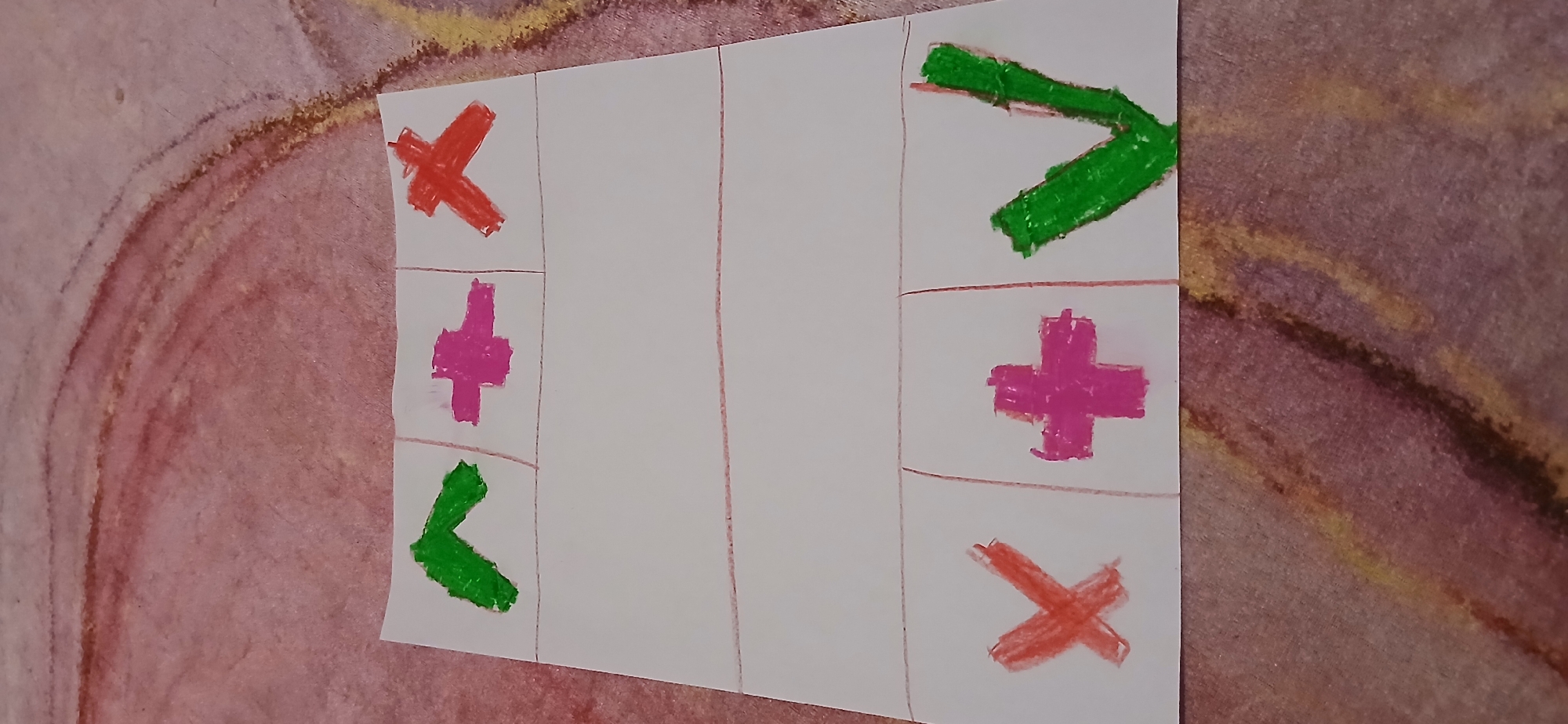 The image shows a piece of white paper with hand-drawn symbols and lines, laid on a textured surface that appears to be a carpet with pink and brown tones. The paper is divided into six rectangular sections by red lines. The top row of sections contains three symbols: a green chevron pointing upwards, a pink plus sign, and a red cross. The bottom row of sections contains three symbols: a red cross, a pink plus sign, and a green checkmark. The bottom row also has red lines dividing the sections. The image does not contain any identifiable landmarks or elements that would allow for its location to be determined within the city of Samarinda, Indonesia.