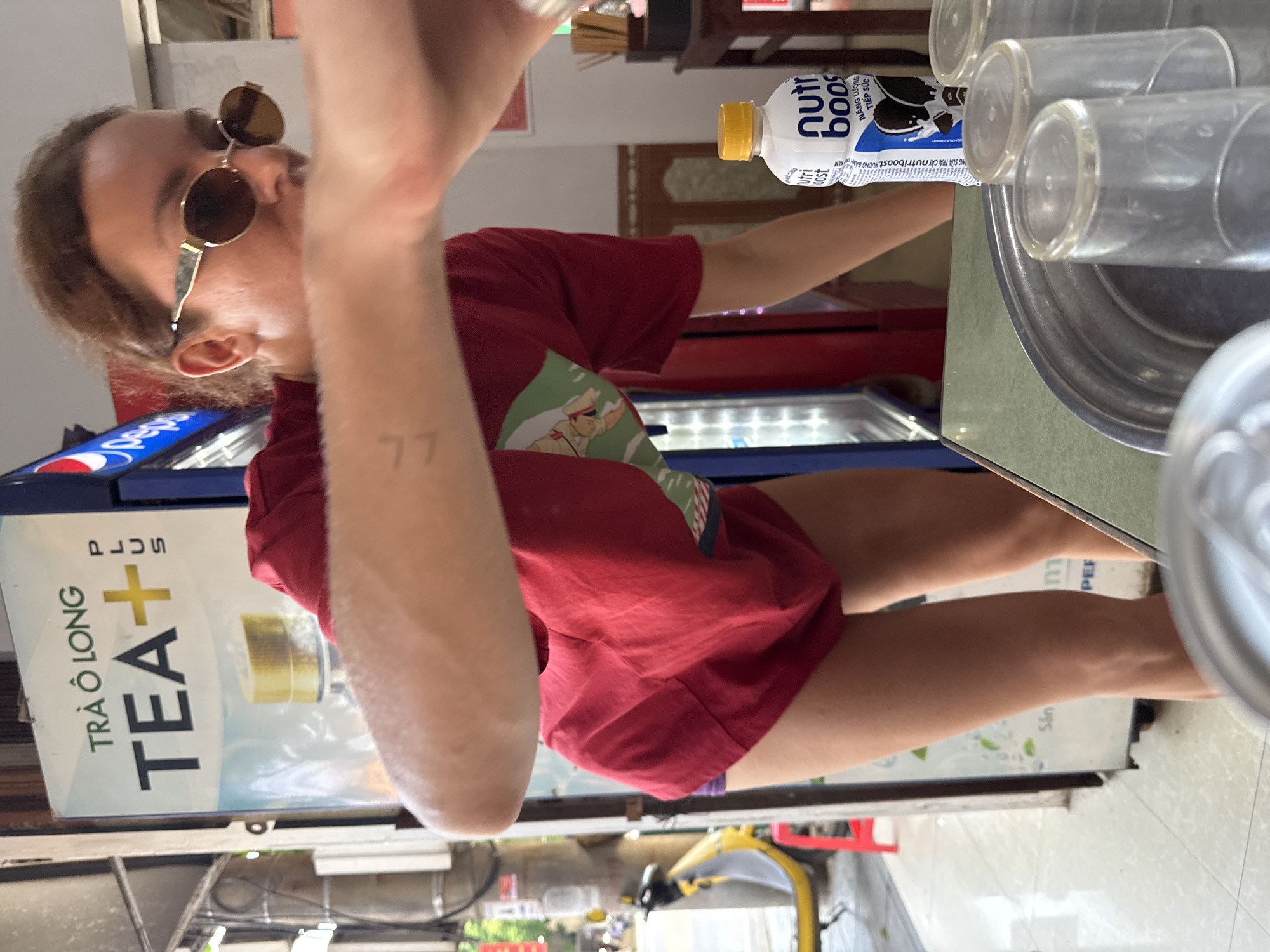 This image, taken in Phố Ràng, Vietnam, depicts a young person in an informal eatery or store setting. The entire composition appears to be rotated ninety degrees clockwise. The central subject, visible from the chest up, has brown hair and wears round, gold-rimmed sunglasses, obscuring their eyes. Their left arm is raised, holding an unseen container to their mouth as if drinking. A clear "77" tattoo is visible on their left forearm. They are dressed in a red t-shirt with a graphic on the front depicting a figure in a hat, possibly a uniform.
To the right of the person, on a counter, sits a white "nutriboost" bottle with a yellow cap, clearly labeled "NĂNG LƯỢNG TIẾP SỨC" (Energy Boost) and "HƯƠNG BÁNH KEM" (Oreo/cookie cream flavor). Several clear plastic cups are stacked on a metallic tray beside the bottle.
In the background, behind the person, a blue cooler with a Pepsi logo is visible. To the left, a display board advertises "TRÀ Ô LONG TEA+ PLUS". The lower right of the frame shows the person's legs, implying they are seated or squatting. A bright, naturally lit street scene with a yellow scooter or motorcycle is visible in the distant background, indicating it is daytime. The various Vietnamese texts on packaging and displays confirm the location. Emotions are not discernible due to the subject's obscured face.