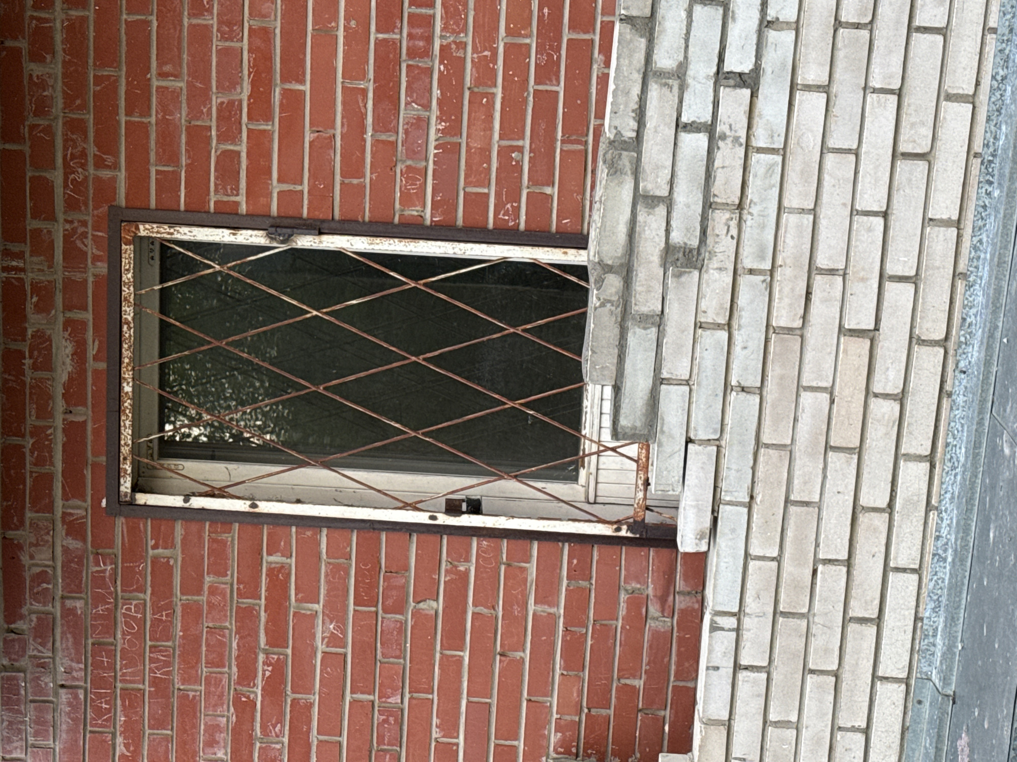 A brick wall, composed of red rectangular bricks, is visible. A window with a metal security grill is set into the wall. The window frame is painted white. To the right of the red brick wall is a light gray brick wall. On the red brick wall below the window, inscriptions or graffiti are present. The scene is located in the city of Unknown, Unknown.