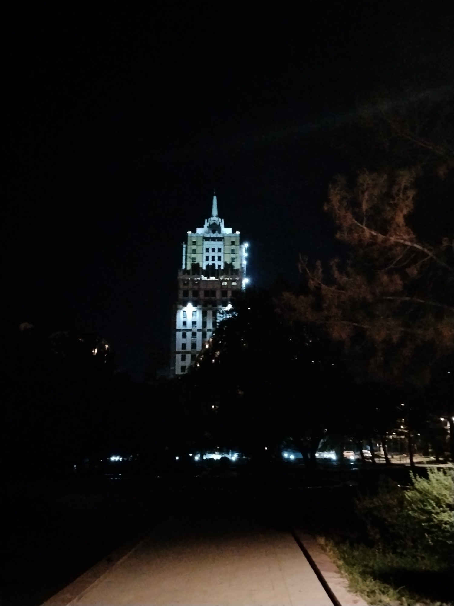 An exterior shot captures a nighttime view of the White House in Bishkek, Kyrgyzstan, illuminated against the dark sky. The imposing building, with its distinctive architecture and spire, stands centrally in the frame. In the foreground, a paved pathway is visible, bordered by dark vegetation and trees, some with reddish-brown foliage. In the middle ground, the building is partially obscured by trees. Distant lights suggest the presence of other structures or streetlights within the city. The overall scene is dimly lit, with the building's lights providing the primary illumination.