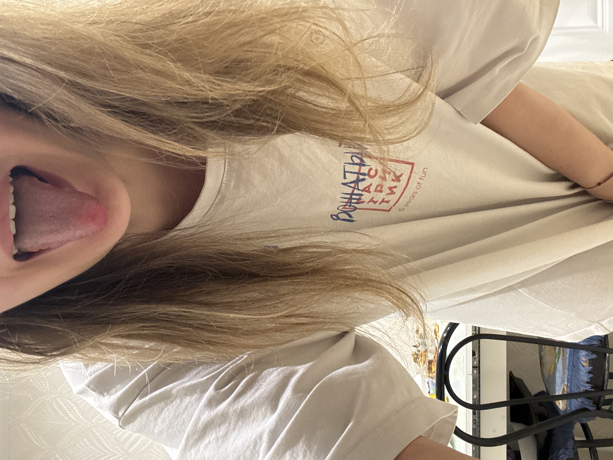 The image shows a fair-skinned person with long, blonde hair sticking out their tongue. They are wearing a light-colored T-shirt with some text in Russian and English that appears to say "5 years of fun". In the background is a metal structure which could be a chair or clothing rack. There is no evidence in the image that indicates a location in the city of Izhevsk, Russia.
