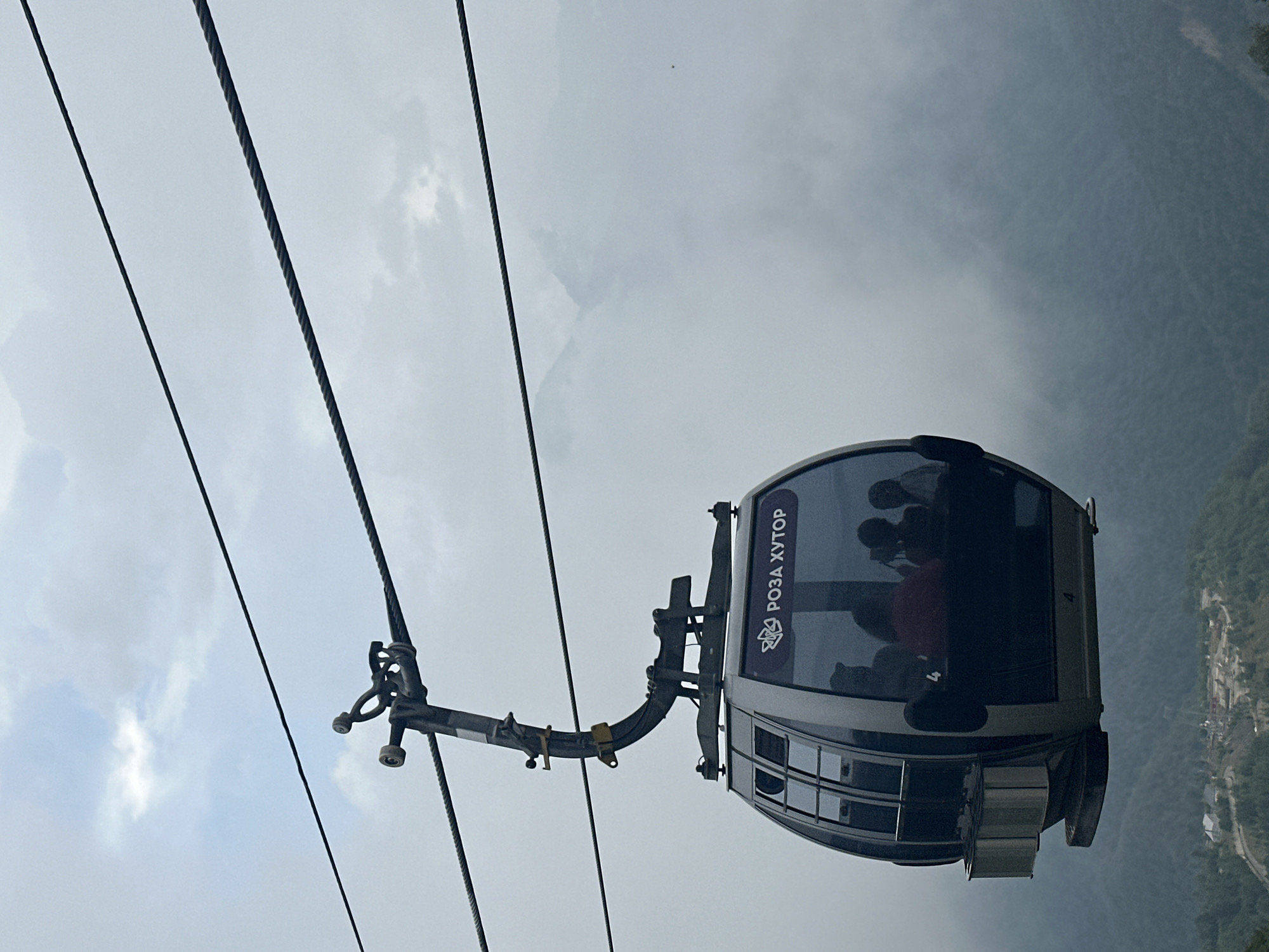 A cable car is descending through a cloudy mountain landscape. The cable car is gray and oval-shaped, with tinted windows. Inside, several people are visible, silhouetted against the light. The text "РОЗА ХУТОР" is written on the side of the cable car in purple lettering. The cable car is suspended from a complex metal mechanism attached to thick cables. Below, a mist-shrouded mountain range is visible, with a small settlement or resort area in the distance. The sky is overcast with clouds. The scene appears to be in the ski resort of Rosa Khutor, located in Estosadok, Russia.