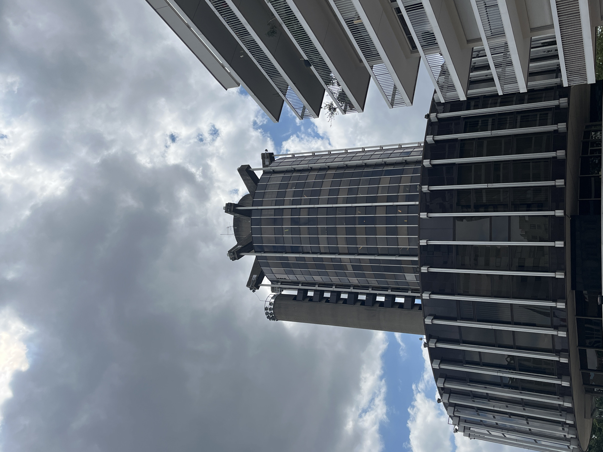 This is a low-angle, outdoor, full-shot photograph looking up at modern buildings against a cloudy sky. The image is oriented vertically. Two distinct architectural styles are visible. On the left and top of the frame, there are the undersides of multiple levels of a building with a modern design featuring cantilevered sections and white railings. Below this, and dominating the center and right of the frame, is a tall, cylindrical skyscraper with a dark, reflective glass facade. The skyscraper has horizontal bands of windows and several protruding sections with external lighting fixtures. A large, cylindrical structure, possibly an elevator shaft or ventilation tower, is attached to the side of the skyscraper. The sky is filled with a mix of dark, heavy clouds and patches of bright blue sky with scattered white clouds, suggesting a dynamic weather condition. The lighting suggests it is daytime, possibly late afternoon or overcast conditions. No people are visible. The scene appears to be an urban setting, likely a business district given the modern architecture. The crops highlight the details of the buildings' facades and the cloud formations in the sky. There is no visible text in the image.