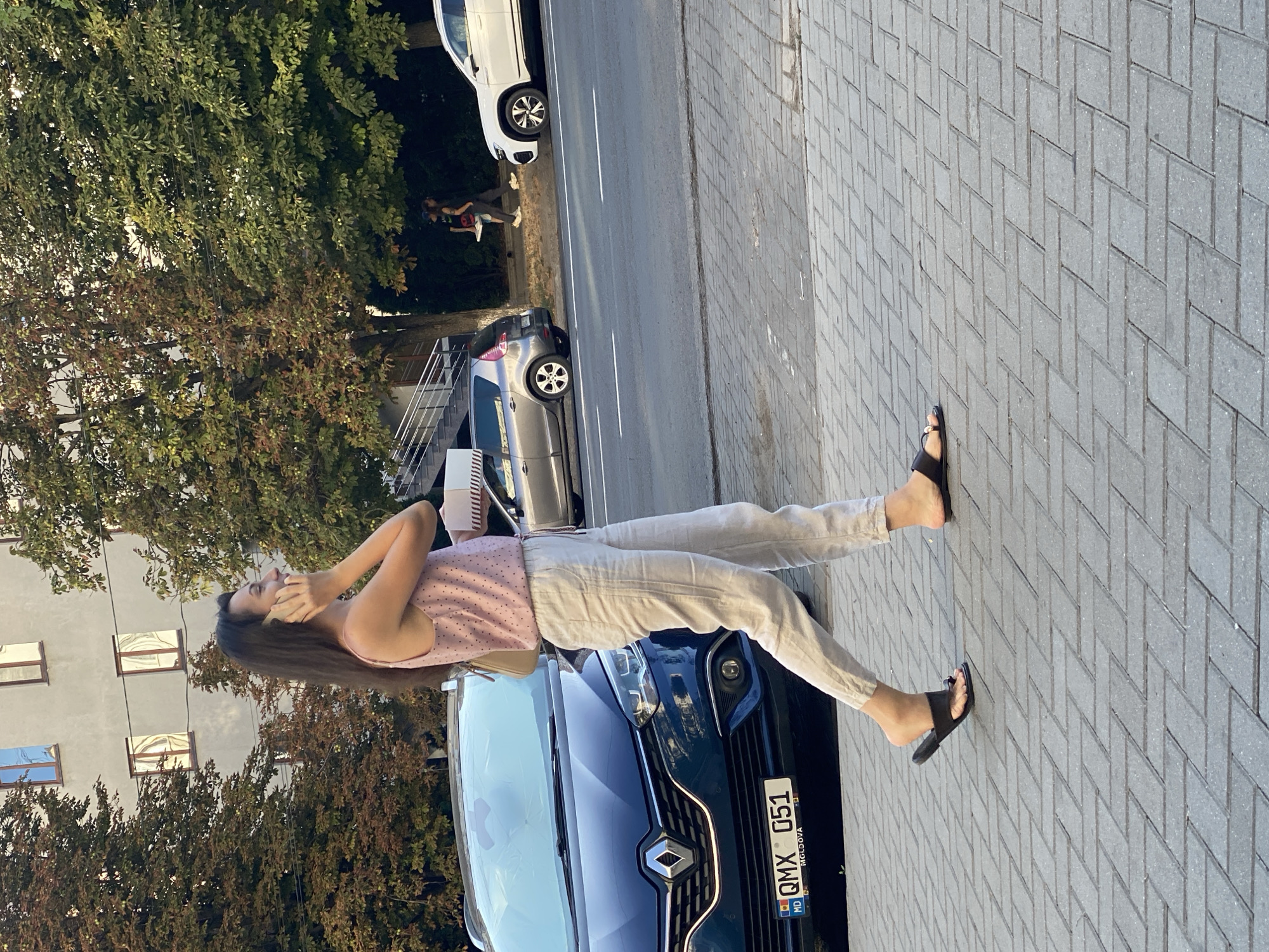 A woman with long, dark hair is walking on a paved sidewalk, talking on a mobile phone. She is wearing a pink polka-dot tank top and loose, light-colored linen pants. She has on black slip-on sandals. In her right hand, she is holding a small, light-colored box, possibly a cake box. She is walking past a dark blue Renault car, which has a Moldovan license plate that reads "MD QMX 051". The car is parked on the side of a street. Further down the street, a silver car is also visible, along with a white SUV. In the background, there is a building with windows reflecting the sky, and trees with green and reddish-brown leaves, suggesting it is autumn. Two people are walking in the background, away from the camera. The overall setting appears to be an urban environment in Chișinău, Moldova, judging by the license plate and common vehicle models.