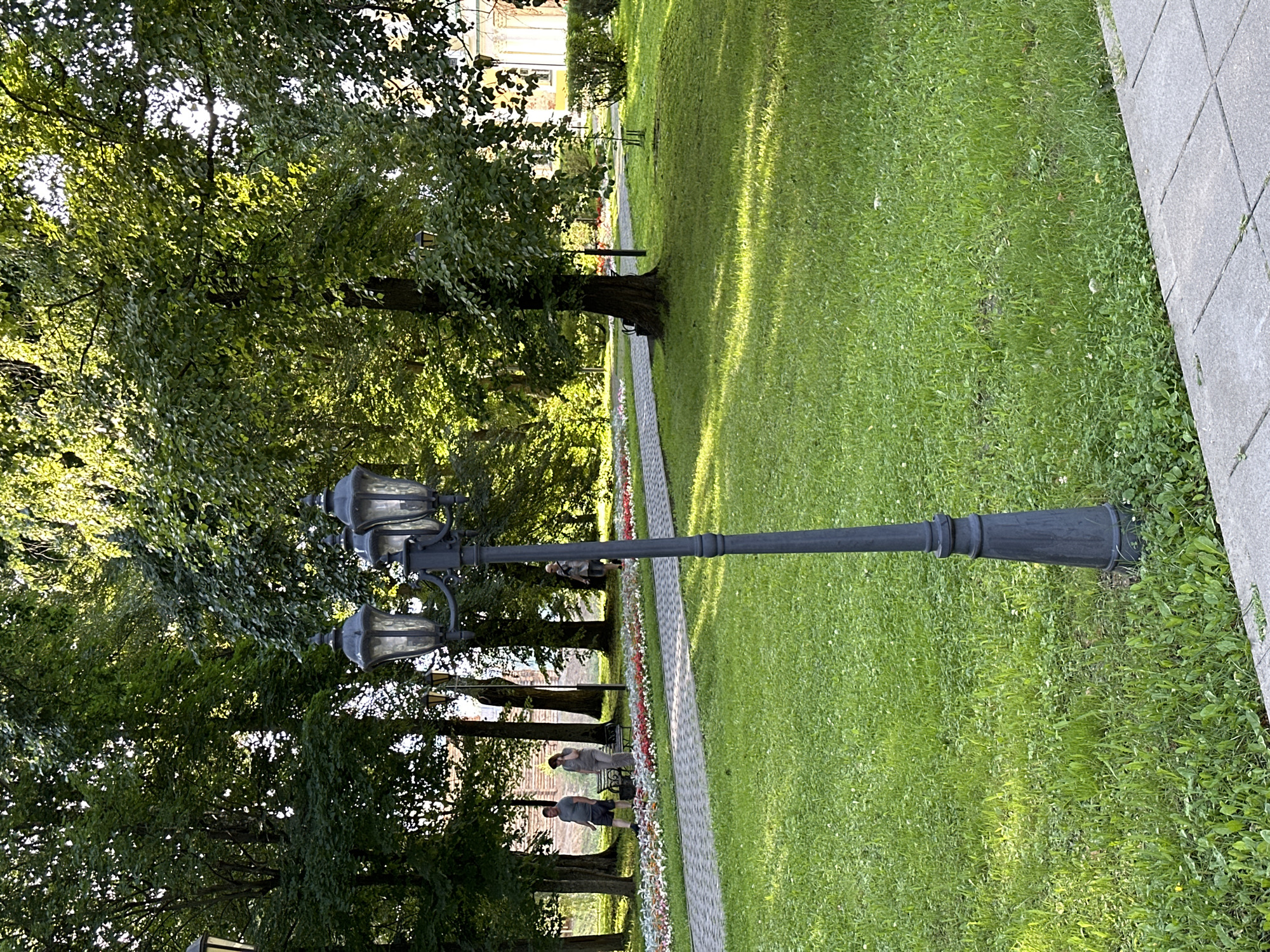 The image depicts a park-like setting in Selivanovo, Russia, on a sunny day. In the foreground, a tall, dark-colored lamppost with three lamp heads stands on a patch of green grass. To the left of the lamppost, a paved walkway runs diagonally across the scene. Adjacent to the walkway is a flowerbed with rows of small, colorful flowers. Beyond the flowerbed, a group of mature trees with dense green foliage forms a backdrop. Two individuals are visible walking on the paved path, with a third person further down the path. Sunlight filters through the leaves of the trees, creating dappled shadows on the grass and path. In the background, a building with light-colored walls and a decorative facade is partially visible through the trees. The scene conveys a peaceful atmosphere typical of a public park in the city.