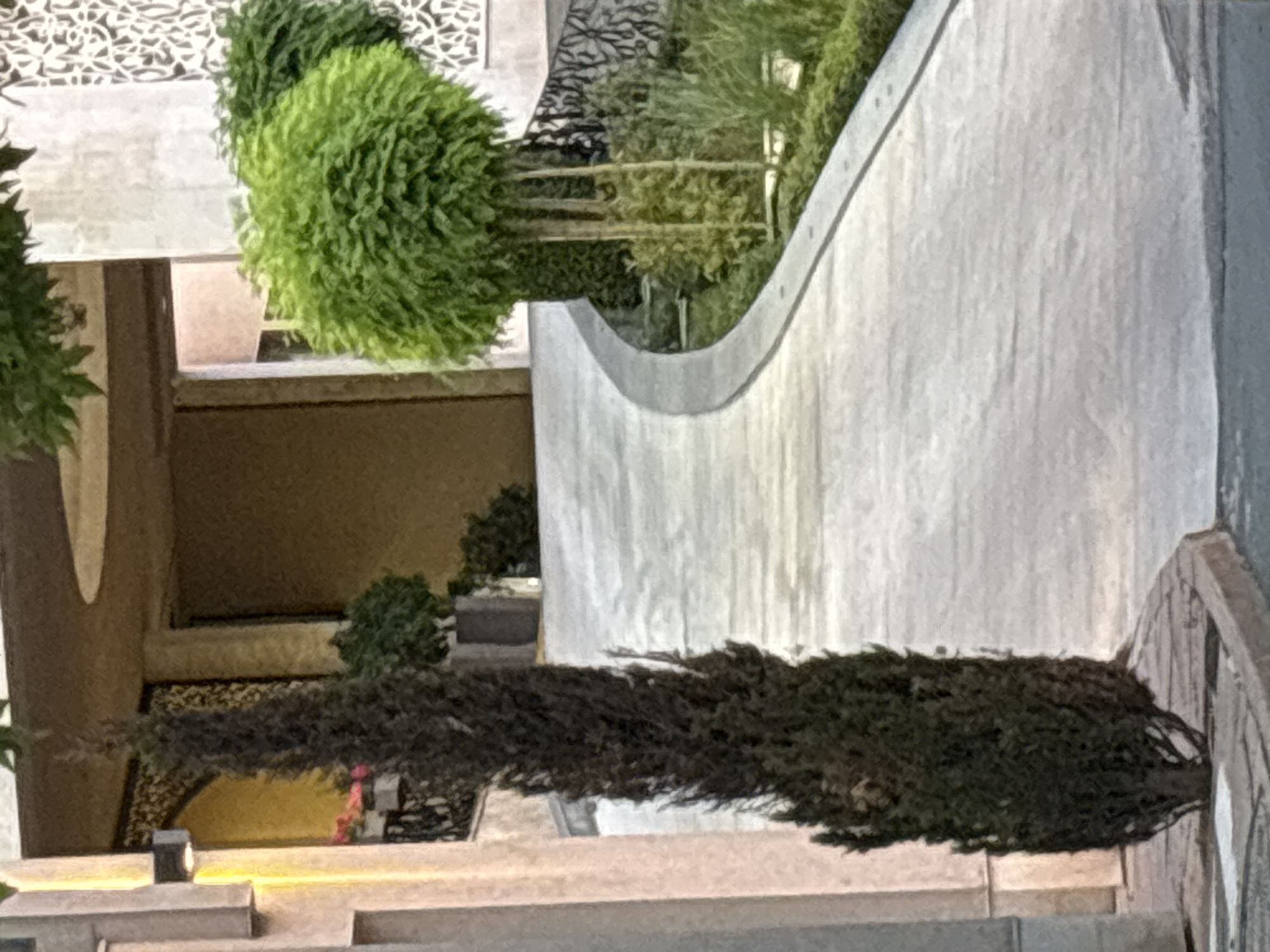 The image depicts a pathway curving through a well-maintained garden area. On the left, a tall, dark green, columnar evergreen tree stands prominently. To the right of the pathway, a vibrant green, rounded shrub is visible, supported by several thin, vertical stakes. The pathway itself appears to be made of concrete and is bordered by lush green foliage and landscaping. In the background, architectural elements of a building are visible, including decorative latticework and a large doorway. The overall scene suggests an exterior view of a landscaped area adjacent to a building, possibly in a residential or commercial setting. No people are present in the image. The image does not provide any specific geographical markers that would allow for the identification of the city of Nesebar, Bulgaria.