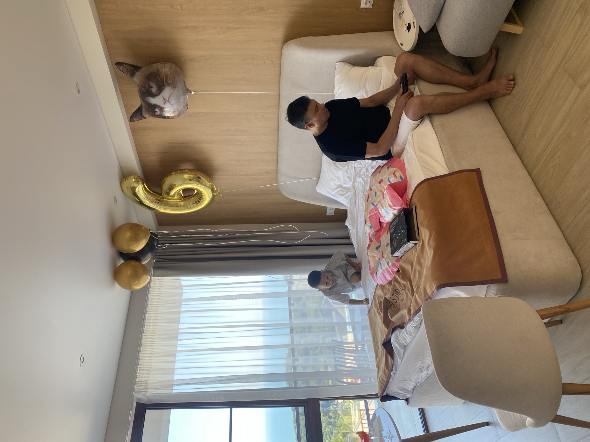 In a hotel room in Dagomys, Russia, a man sits on the edge of a bed playing a video game. Beside him, a young boy crawls on the bed. The room is decorated with balloons, including a "6" shaped balloon and a cat-shaped balloon. A large window offers a view of the ocean and surrounding greenery.