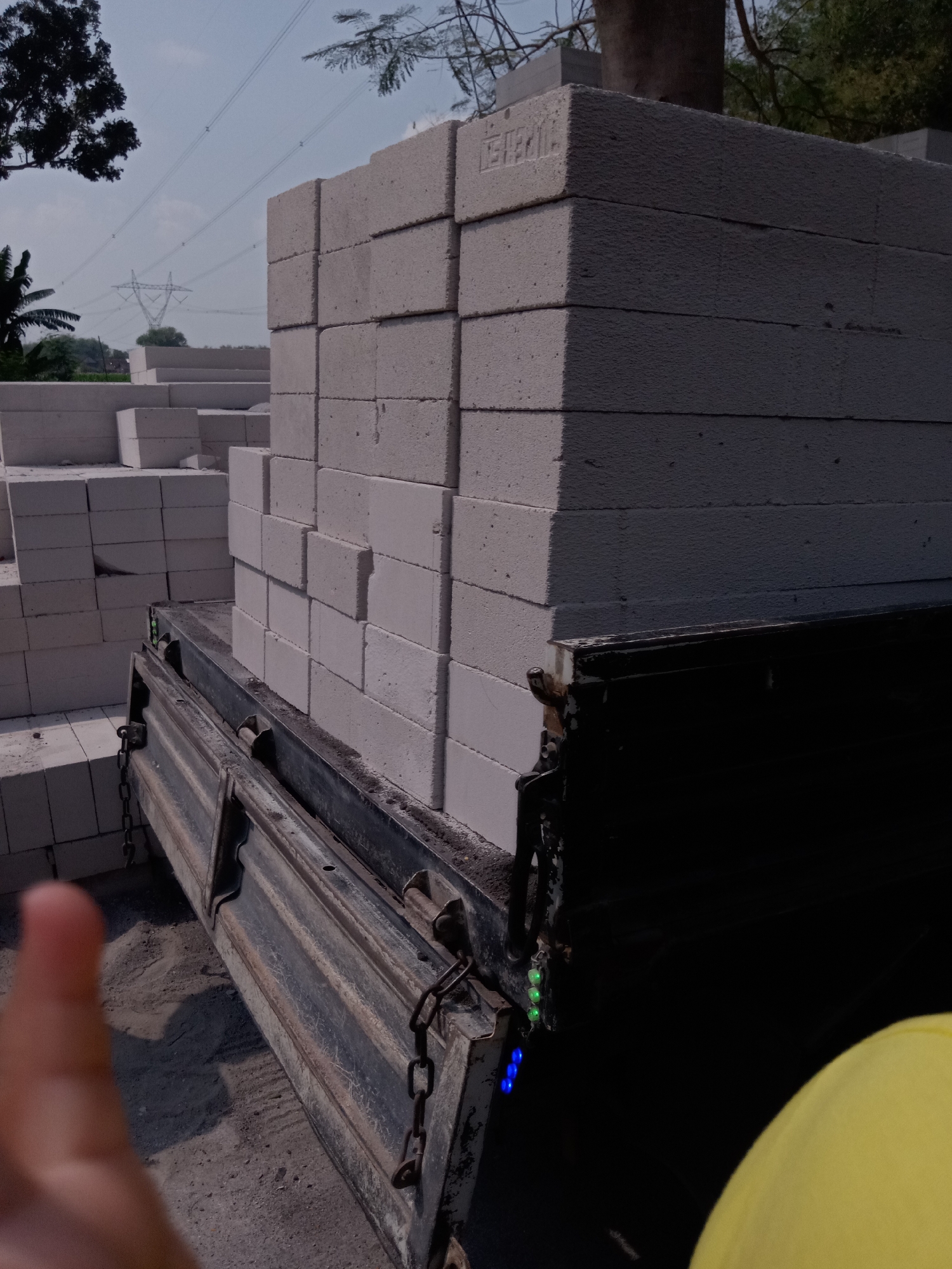 The image captures a scene in Sugihwaras, Indonesia, focusing on a truck bed loaded with stacks of lightweight, porous concrete blocks. The blocks are light gray in color and appear to be of a uniform size, stacked neatly. In the background, a large electrical transmission tower and power lines are visible against a hazy sky, suggesting an outdoor, possibly industrial or construction-related setting. Some greenery, including trees and branches, frames the upper portion of the image. The truck bed itself is dark, showing signs of wear and rust, with a chain attached to its side. In the foreground, a blurred thumb is visible, indicating a human presence, and a yellow object, possibly clothing, is partially visible in the bottom right corner. The ground appears to be covered in sand or loose soil.