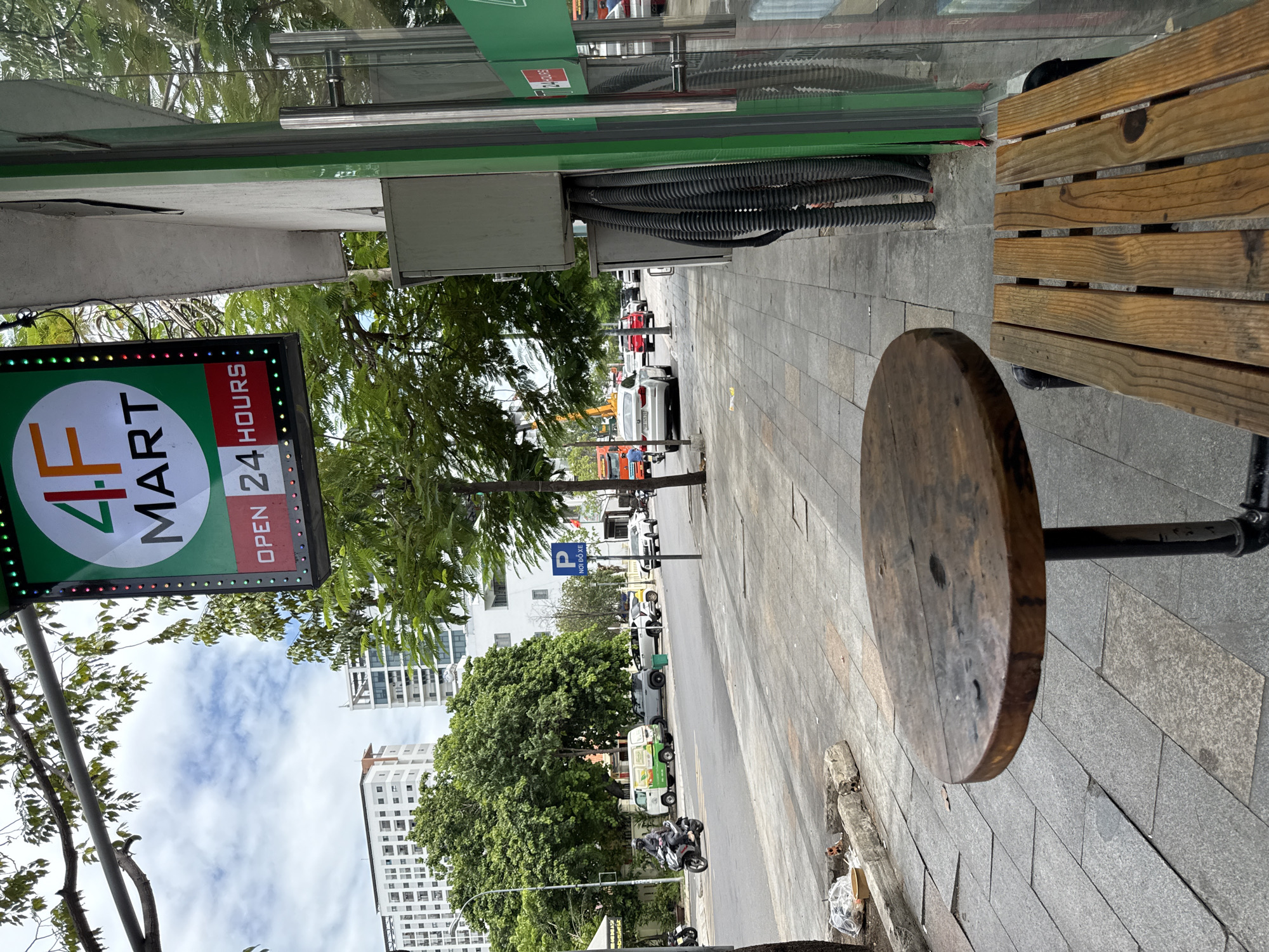 This image depicts a street scene in Nha Trang, Vietnam, with a focus on the exterior of a "4F MART" convenience store. In the foreground, a rustic wooden table and a matching bench are positioned on a grey tiled sidewalk. Beyond the seating area, a street runs horizontally across the frame, with vehicles including a scooter, a white van, a grey SUV, a yellow car, and several white cars driving or parked. A blue "P NOIDO XE" (parking) sign is visible on the right side of the street. The "4F MART" store's facade features a large green sign with a white circle containing "4F" in red and orange, and "MART" in black, along with "OPEN 24 HOURS" in red text. The store has a glass door with silver handles and a green frame. Trees and buildings, including modern high-rises, are visible in the background, suggesting an urban environment. The sky is partly cloudy.