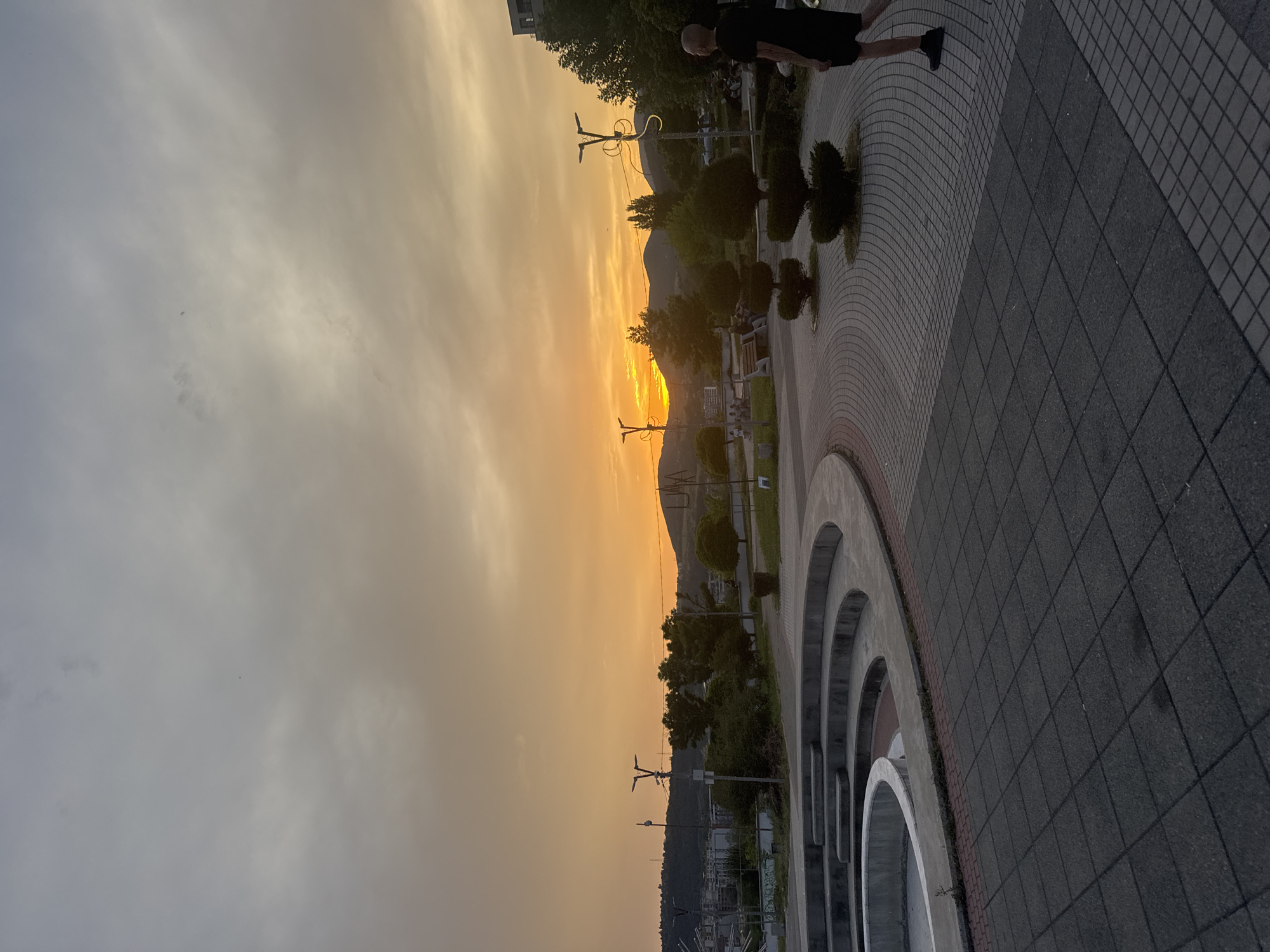 A wide shot captures an outdoor urban plaza at sunset. The sky is dominated by a vibrant orange glow emanating from the sun setting behind a range of hills on the horizon. Clouds are scattered across the sky, tinged with the sunset's colors. In the foreground, a circular, tiered concrete structure, possibly a fountain base, is visible on the left. The ground is paved with dark square tiles arranged in a grid pattern.
To the right of the fountain structure, a person with a shaved head, wearing a black t-shirt and dark shorts, walks away from the camera. The plaza features several manicured, topiary-style bushes and lampposts. In the middle ground, there are benches and more trees, with a few people seated on the benches and walking. Beyond the plaza, a city landscape with buildings and more hills is visible against the colorful sky. The overall impression is of a serene evening in a public urban space.