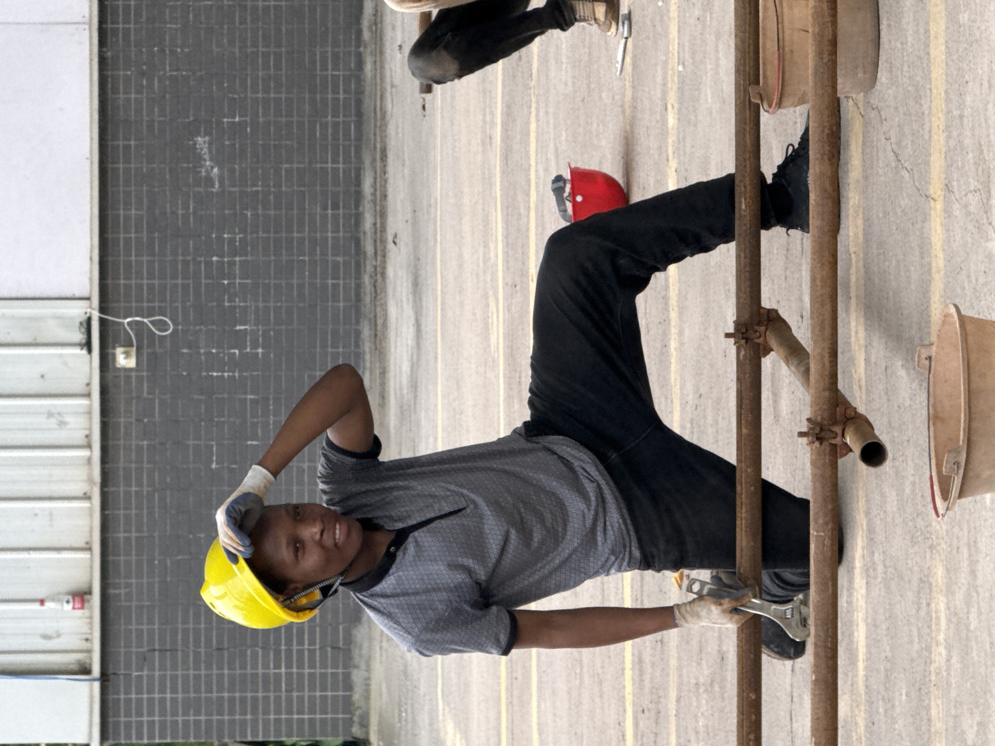 In Jiangjin, China, a young construction worker, wearing a yellow hard hat and a grey collared shirt, kneels on a paved surface marked with yellow lines. The worker is holding a wrench in their right hand, appearing to adjust a metal scaffolding pipe. The worker's left hand is raised, touching the brim of their hard hat. Black pants and work gloves are visible. In the background, a grey brick wall and a white structure are present. Red and tan buckets are also scattered around the work area.