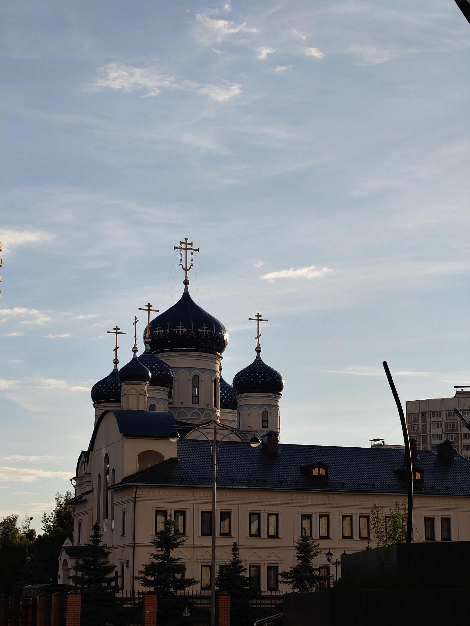 This image captures an Orthodox church in Moscow, Russia, under the warm glow of the golden hour. The central subject is a multi-domed church building, featuring several dark blue, almost black, onion domes topped with golden Orthodox crosses. The largest cross, on the prominent central dome, includes an anchor-like detail at its base. The church's main structure is a light beige or cream color, with contrasting dark roofs on its lower sections. Architectural details like arched windows and decorative moldings are visible on the building and the drums supporting the domes.
In the foreground, small evergreen trees line a dark fence. A dark lamppost or utility pole stands on the right, partially obscuring a multi-story, light-colored residential building in the background. A faint structure is also visible on the far left edge.
The sky is a clear, pale blue, with scattered white, wispy clouds. The low-angle sunlight illuminates the church domes and crosses from the left, creating bright highlights and emphasizing the architectural forms. There are no people or discernible activities taking place, and no visible text in the image. The scene evokes a sense of peaceful grandeur.