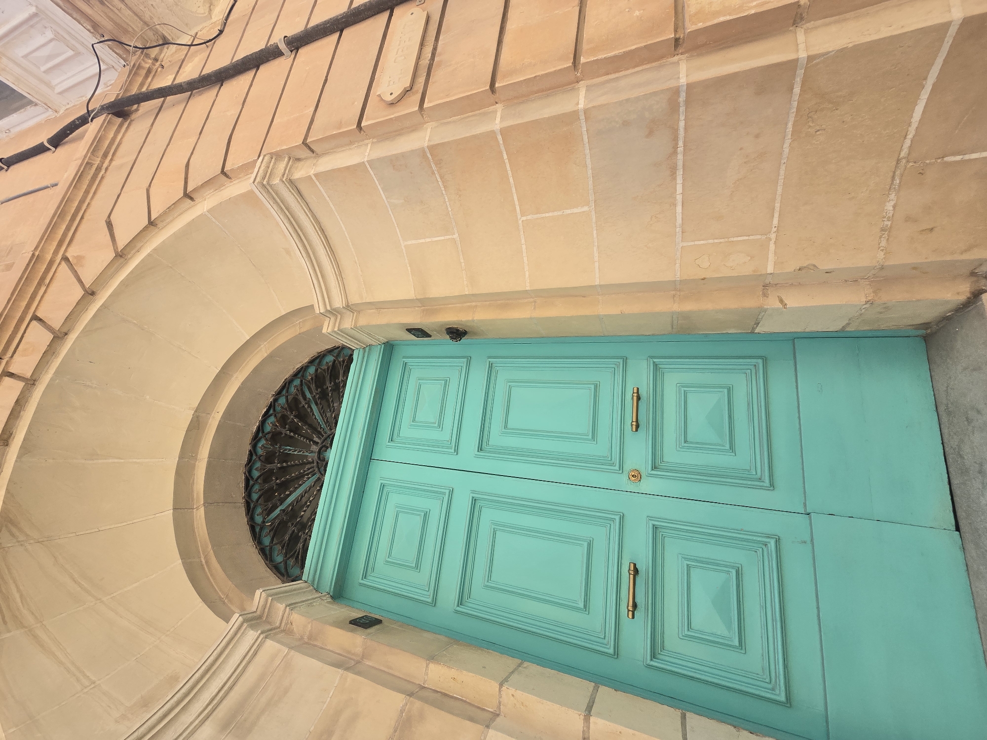 This is a low-angle, eye-level, outdoor, full shot of a bright turquoise double door with ornate metalwork above it, set within a stone archway on a building in Sliema, Malta. The doors are paneled with recessed rectangular sections and feature brass handles and a central lock. The stone facade of the building is constructed from large, light-brown blocks with white mortar lines. A street sign reading "TRIQ OROPA" is visible on the wall to the right of the door. Electrical conduits run vertically up the side of the building. The lighting suggests it is daytime.