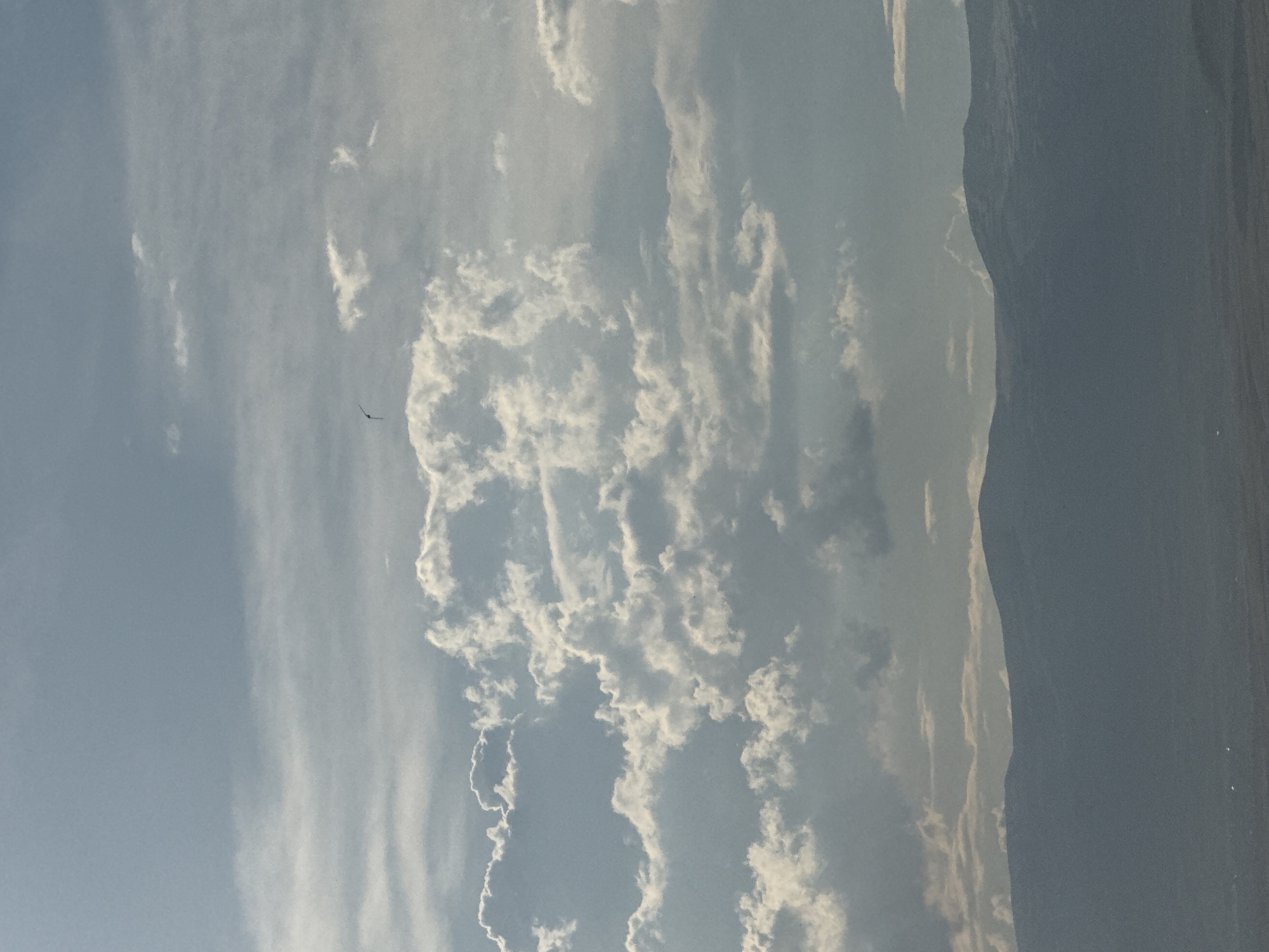 The image shows a wide shot of the sky above Abovyan, Armenia. The sky is filled with a mix of puffy white clouds and thinner, wispy clouds against a pale blue backdrop. A single dark bird is visible in flight high in the sky. Below the sky, a silhouetted mountain range stretches across the horizon. The lower portion of the image shows a dark, hazy landscape beneath the mountains.