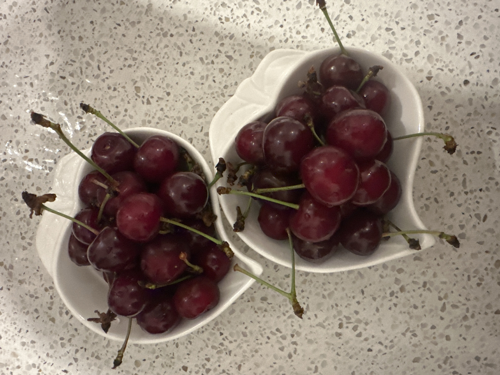 Two white heart-shaped bowls are filled with dark red cherries. The bowls are placed on a speckled, light-colored countertop. The cherries have green stems, some of which are detached. The image is taken from a top-down perspective. The scene is indoors.