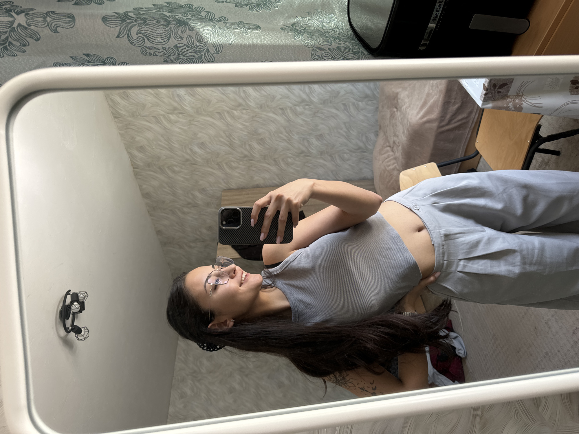 The image captures a mirror selfie taken by a young woman in an indoor setting. The woman, with long dark hair and wearing clear-framed glasses, is seen from the chest up, smiling slightly as she holds a smartphone with a textured black case to take the picture. She is dressed in a light gray ribbed crop top and light gray pleated wide-leg trousers, revealing a portion of her midriff. A tattoo featuring celestial elements like a crescent moon and stars is visible on her left arm.
The mirror reflects various elements of the room. A wall with a textured, possibly faux fur or shaggy, white covering is prominent, along with another wall featuring a more abstract, wavy pattern. In the background, a bed or couch is visible with a patterned duvet or blanket, featuring a gray-green floral or leafy design. A dark-colored appliance, clearly branded "Tefal," rests on a table covered with a light-colored, patterned tablecloth. A simple wooden chair is also visible near the table. On a wall to the left, a black wall-mounted light fixture with two industrial-style cage lamps is attached. The overall lighting suggests it is daytime. The location context provided is Sofia, Bulgaria, though no specific visual cues within the image confirm this.