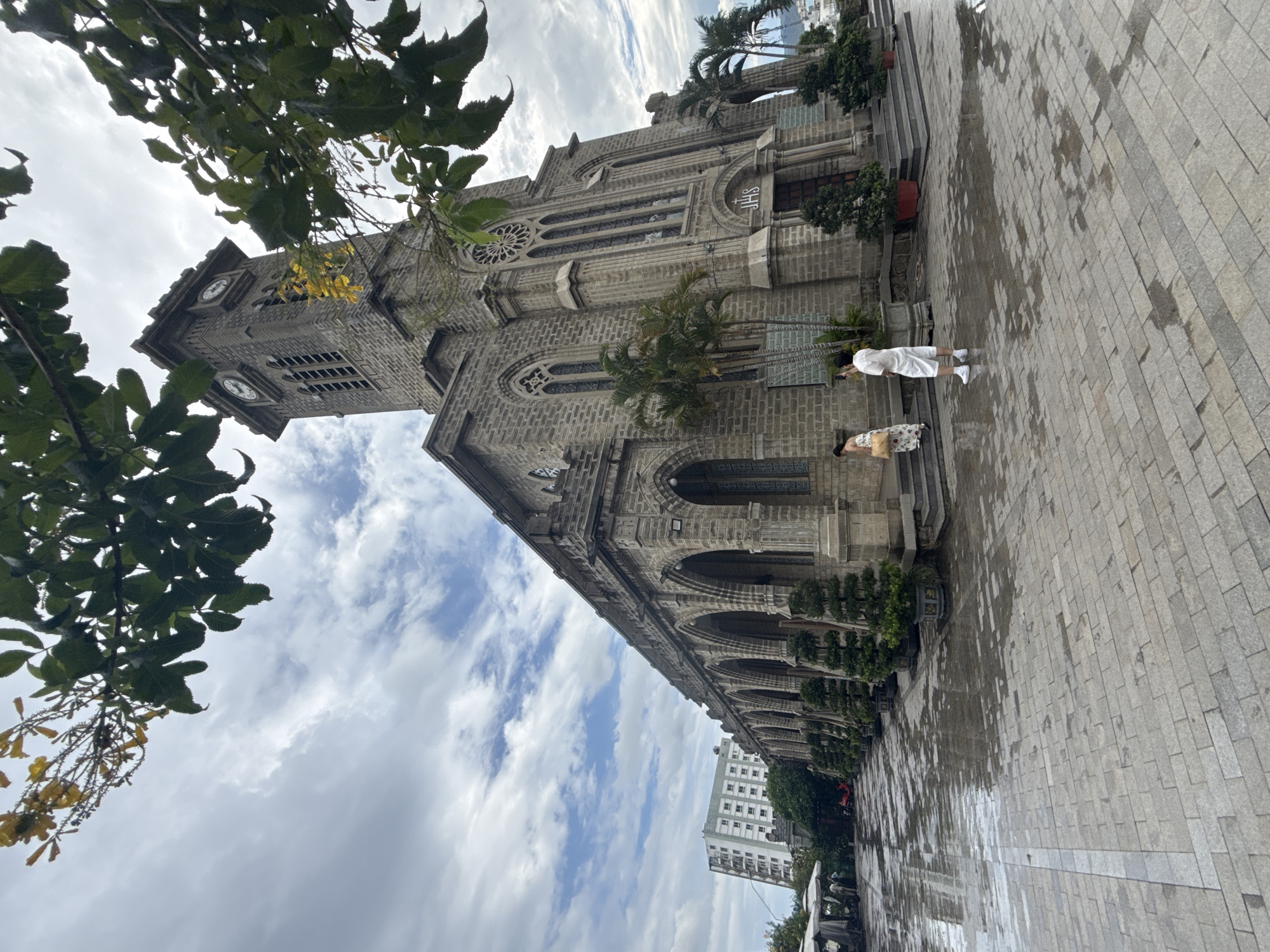 The image shows the Nha Trang Cathedral, a Roman Catholic cathedral in Nha Trang, Vietnam. The cathedral is made of stone and has a tall bell tower with clocks. In front of the cathedral, there is a paved courtyard with reflections of the sky and surrounding structures due to the wetness. Two people are visible on the steps of the cathedral, one in a white outfit and the other in a patterned dress, appearing to be taking pictures. Several potted trees are placed along the side of the building. In the background, a modern high-rise building can be seen, contrasting with the historic architecture of the cathedral. The sky is partly cloudy, with some areas of blue sky visible through the clouds.