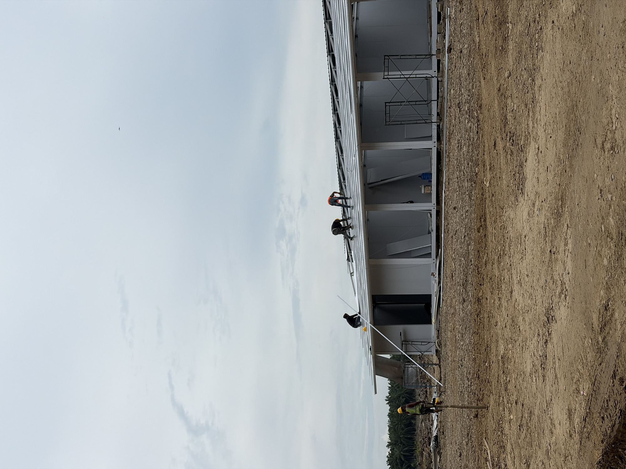 The image shows a construction site in Simpangempat, Indonesia, featuring a long, multi-unit building in the process of being built. The building has a metal roof structure where several workers are present. One worker is wearing a safety vest and helmet and appears to be working on the roof near the edge of the building. Two workers are also on the roof, wearing safety vests. Another worker is on the ground, wearing a yellow helmet and safety vest, possibly supporting or working with materials for the construction. Scaffolding is visible along the side of the building. The ground adjacent to the building is dirt and soil. The sky is overcast. Palm trees are visible in the background.