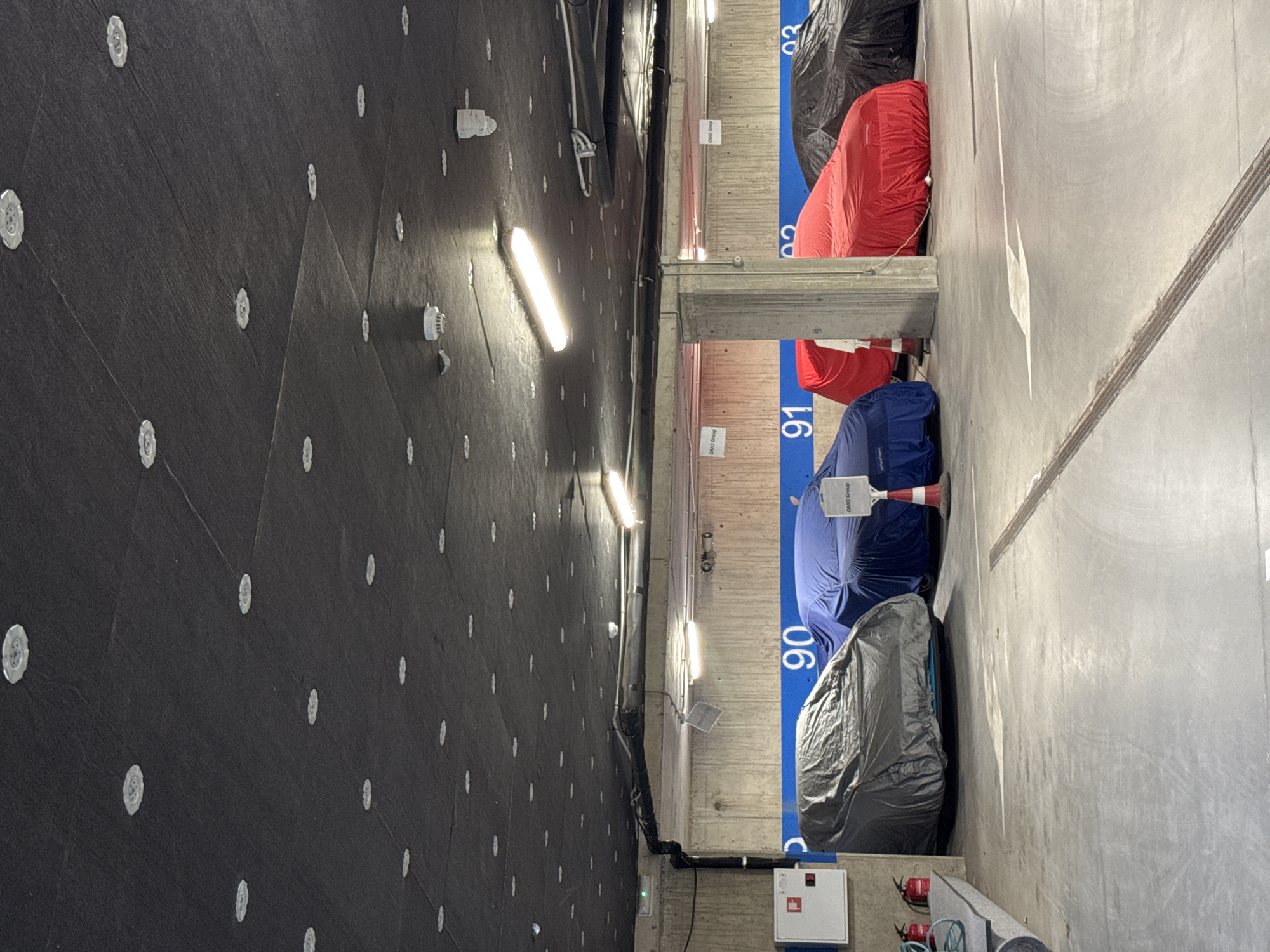 The image depicts an indoor parking garage in Unknown, Unknown. The garage has a dark, textured wall on the left with evenly spaced white circular fixtures. Fluorescent lights run along the top edge of the wall. Parking spots are marked with numbers 90, 91, and 93. Some vehicles are covered with red, blue, and black car covers. A white box with red switches and some rolled-up materials are also visible.