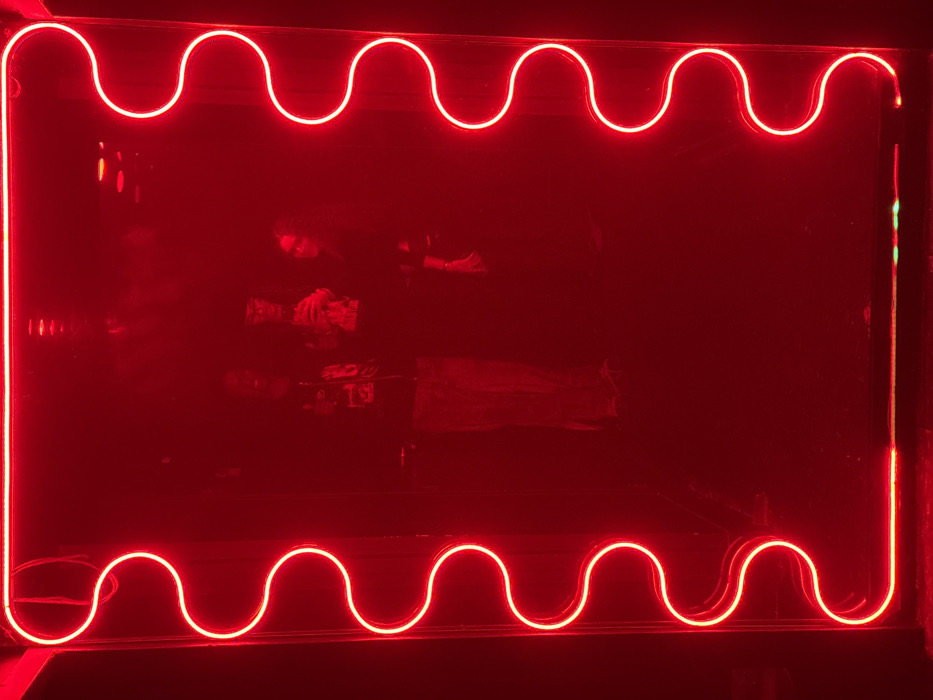 The image displays a dimly lit interior space, saturated in red light. A prominent, bright red neon light fixture forms an irregular, wavy rectangular frame that occupies a significant portion of the visual field. Behind this neon frame, a dark, reflective surface reveals the indistinct reflections of three adult individuals. The leftmost figure is seen wearing dark trousers and a dark top, possibly with a light-colored graphic. The central figure, also in dark clothing, appears to hold an object, possibly a drink glass or phone, near their chest. The rightmost figure wears dark attire and is positioned to the right within the reflection. The environment surrounding the neon and figures is uniformly dark red, indicating low ambient light and the strong luminous output of the neon. The specific location within Paris, France, is not visually identifiable from the image.