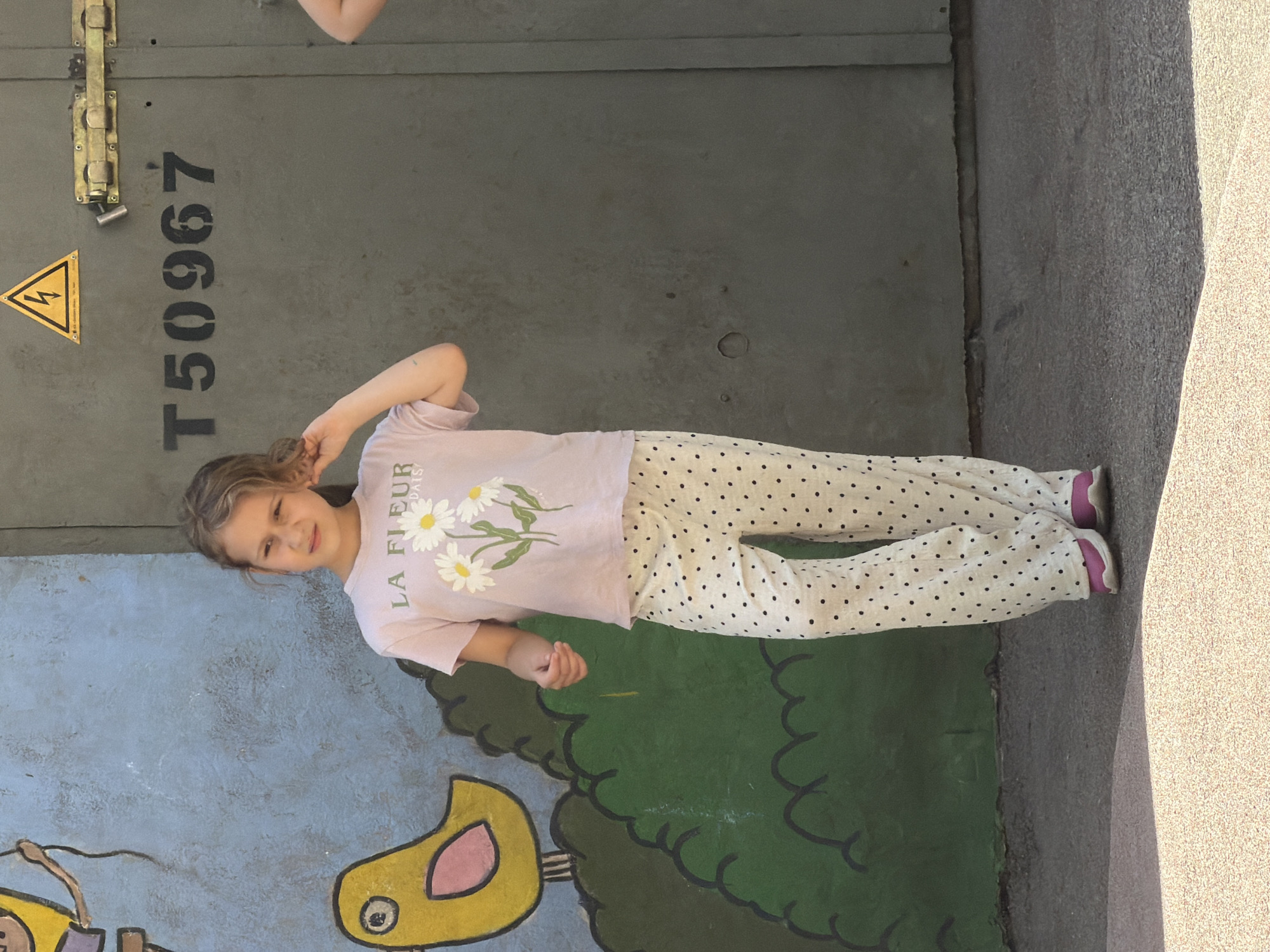 A young girl with blonde hair is standing in front of a grey metal surface that has a yellow lightning bolt symbol in a triangle and the text "T50967" printed vertically on it. The girl is wearing a light purple t-shirt with a daisy print and the words "LA FLEUR" visible, and white pants with black polka dots. She has her right hand near her ear and her left arm is bent at the elbow. Her facial expression is a grimace. In the background, a mural is visible, featuring a blue sky, green bushes, and a yellow bird-like character. The scene appears to be outdoors, possibly on a sunny day given the lighting. The girl's attire suggests it's likely warm weather. The overall impression is of a candid moment captured outdoors, perhaps at a playground or a similar recreational area in Riga, Latvia.
