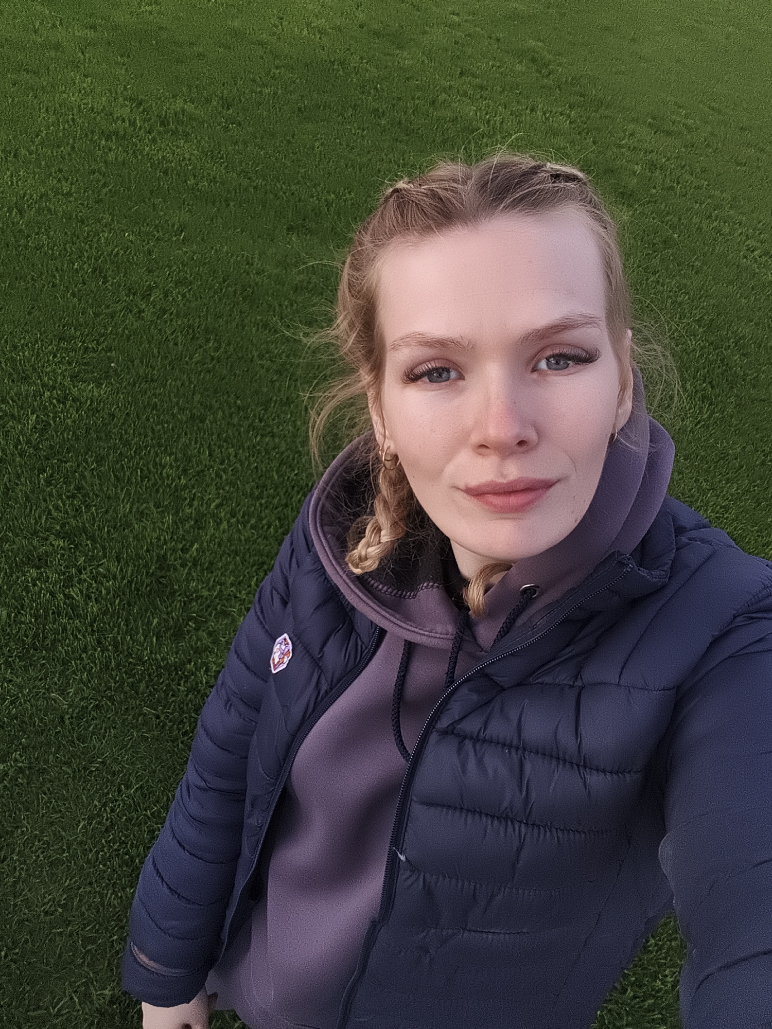 A young woman with blonde hair styled in braids stands outside on a grassy area. She wears a purple hoodie underneath a dark blue puffer jacket. The grass serves as the backdrop for the photo. No other elements suggest the location being Русский Вожой, Russia.