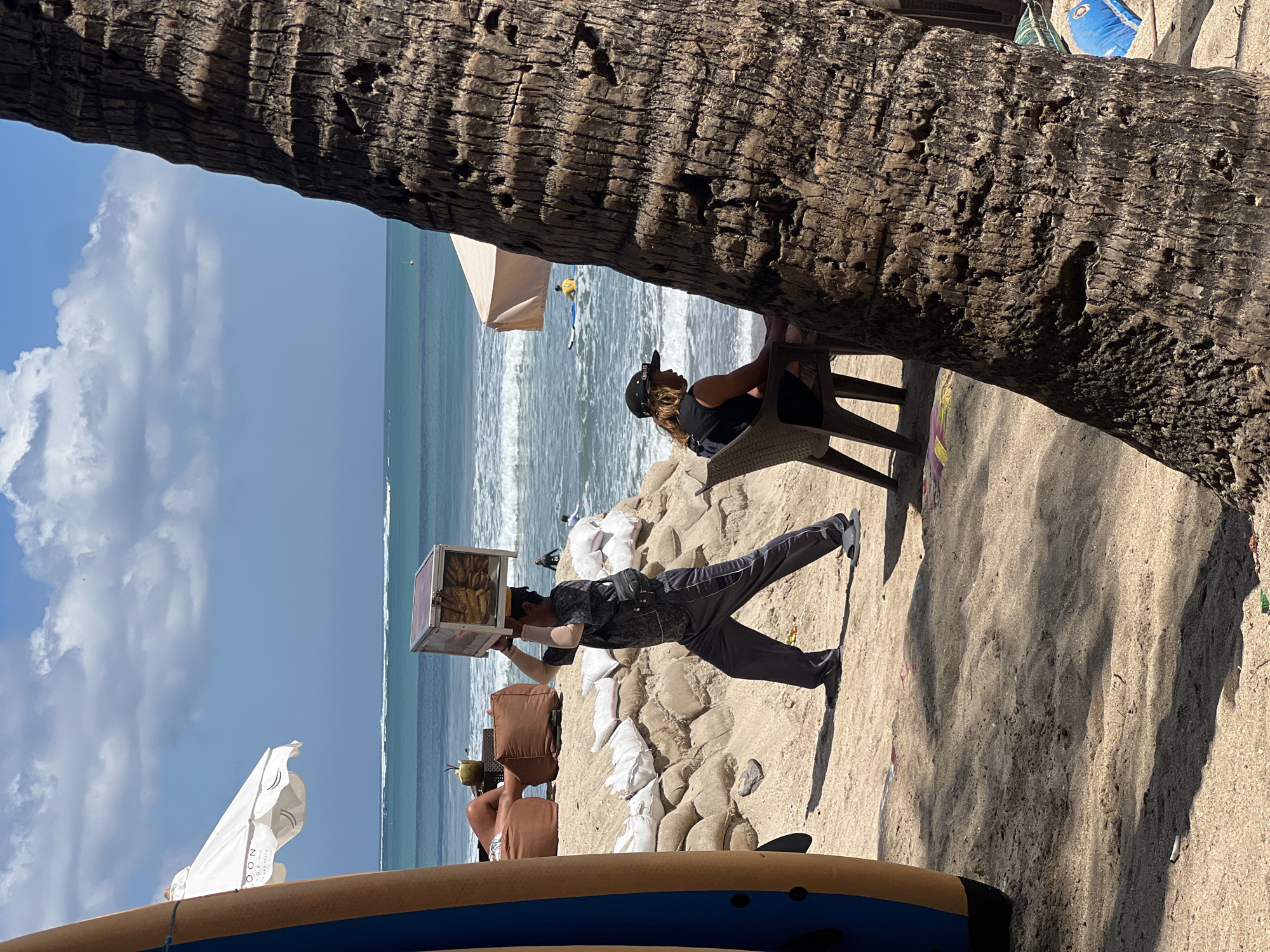 The image captures a sunny day on a beach in Kuta, Indonesia, likely near a beachfront establishment given the presence of lounge chairs and umbrellas. In the foreground, a surfboard stands upright on the left, partially obscuring a large palm tree trunk on the right. A man is walking across the sand carrying a transparent, rectangular box filled with goods on his head. He is wearing dark pants and a dark t-shirt, with a black bag slung across his chest. Behind him, a person is seated at a plastic chair facing the ocean, wearing a black t-shirt and a baseball cap. The beach is lined with sandbags, creating a raised embankment. The ocean in the background has moderate waves, with a few people visible in the water, including one on a surfboard. A white beach umbrella with visible text is also present. The overall scene depicts a bustling beach environment with individuals engaged in various activities.
