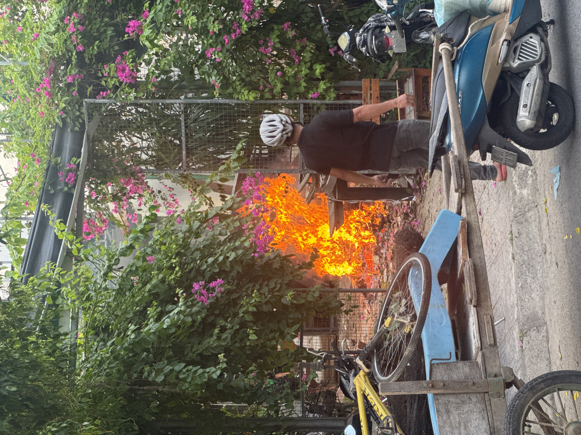 A man in a white helmet and dark clothing tends a burning pile behind a metal fence near a sidewalk. The fire is bright orange and appears to be consuming debris. Green foliage with pink flowers surrounds the scene. A yellow bicycle and a blue and tan scooter are parked nearby. The location is potentially Đà Nẵng, Vietnam, given the common presence of scooters and vegetation in the area. The scene suggests waste disposal or controlled burning activities.