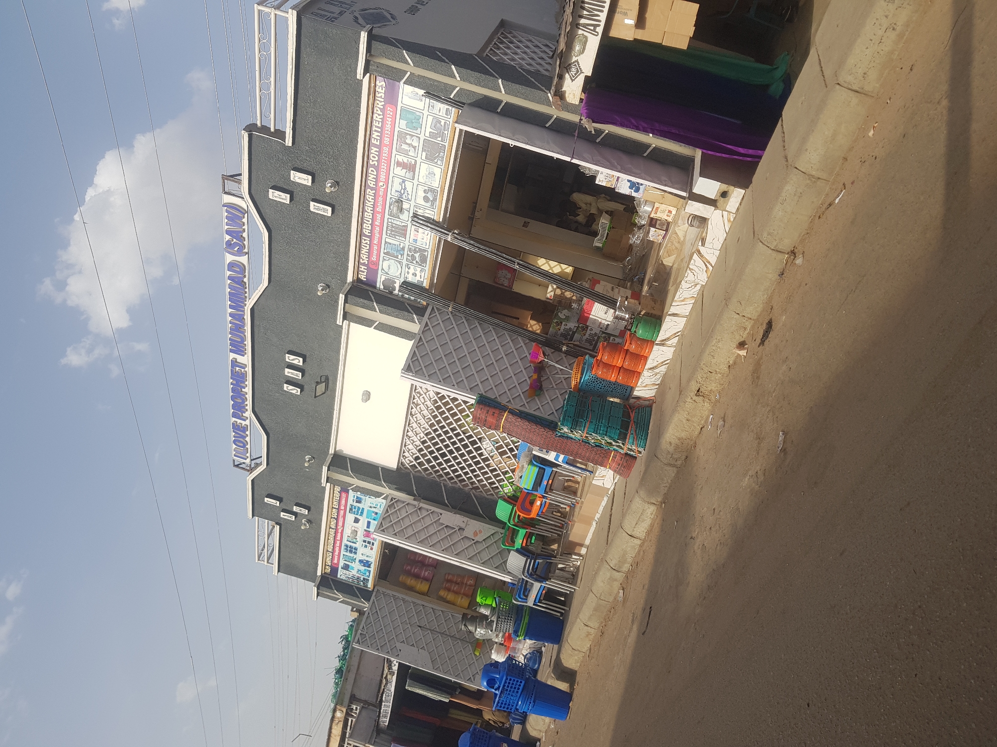 The image shows a street view of a commercial building in Nigeria. The building has two visible storefronts, both appearing to belong to the same business: "ALH SANUSI ABUBAKAR AND SON ENTERPRISES." Merchandise, including plastic containers, baskets, and chairs, are displayed outside on the sidewalk. A sign above the building reads "I LOVE PROPHET MUHAMMAD (SAW)." The location is General Hospital Road, Dutsin-ma, Nigeria. The general environment consists of a street and a sky with some clouds.