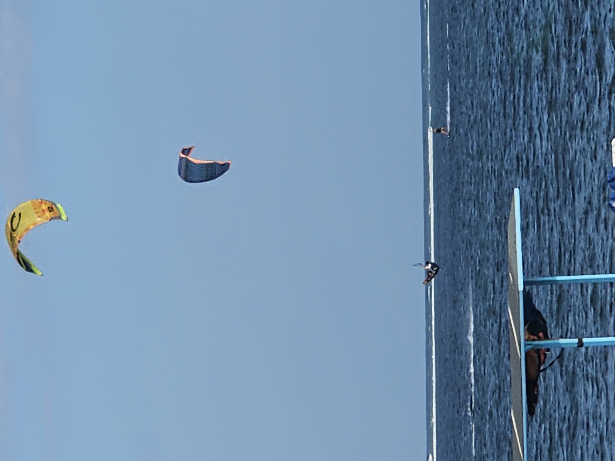 The image shows a clear blue sky over the ocean with several kite surfers. In the upper portion of the image, a yellow and green kite is visible, and below it, a blue and orange kite. Further down, a kite surfer is in mid-air performing a trick. Closer to the water's surface, another kite surfer is riding on the water. In the foreground, a blue and black structure, possibly part of a beachside setup or a floating platform, partially obscures the view of the water and the kite surfers. The ocean is dark blue with some whitecaps indicating waves, and a thin white line of breaking waves is visible on the horizon. The scene appears to be taking place on a sunny day. The location is identified as Denpasar, Indonesia.