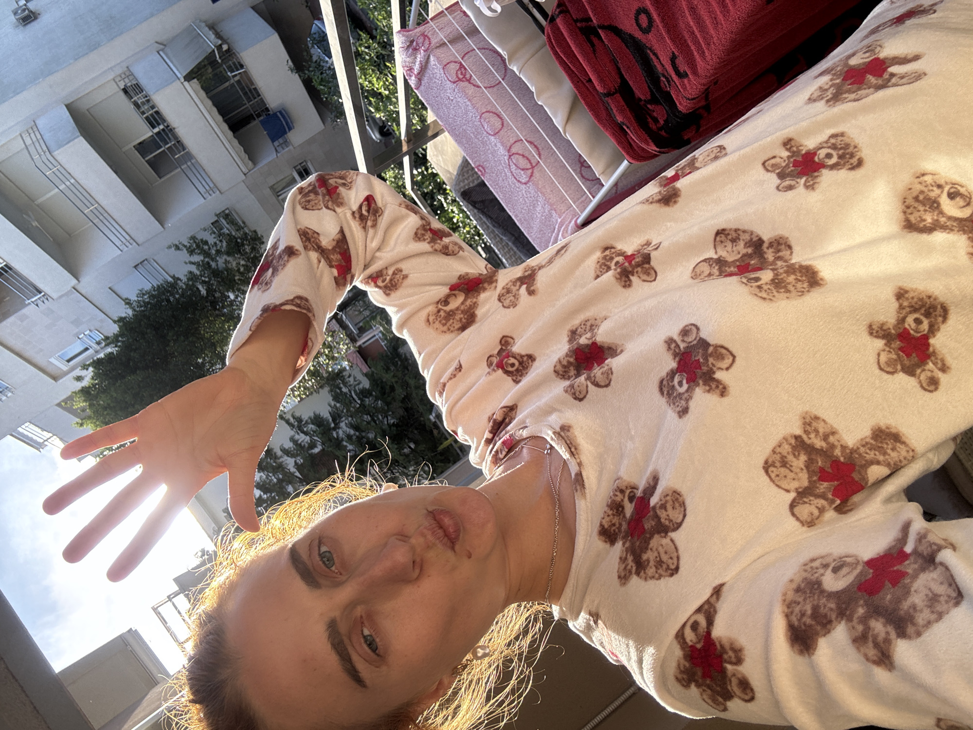 A woman is taking a selfie outdoors in Budva, Montenegro. She is wearing a long-sleeved, off-white pajama top with a teddy bear print. Her left hand is outstretched towards the camera, obscuring the sun, which creates a lens flare. The woman has fair skin, blue eyes, and her blonde hair is sunlit, creating a bright glow around her face.
Behind the woman, there is a clothesline with various items hanging on it, including a pink and white patterned blanket and a red and black patterned blanket. In the background, there are buildings and trees. The buildings appear to be residential, with white walls and balconies. The sky is visible in the upper left corner of the frame.