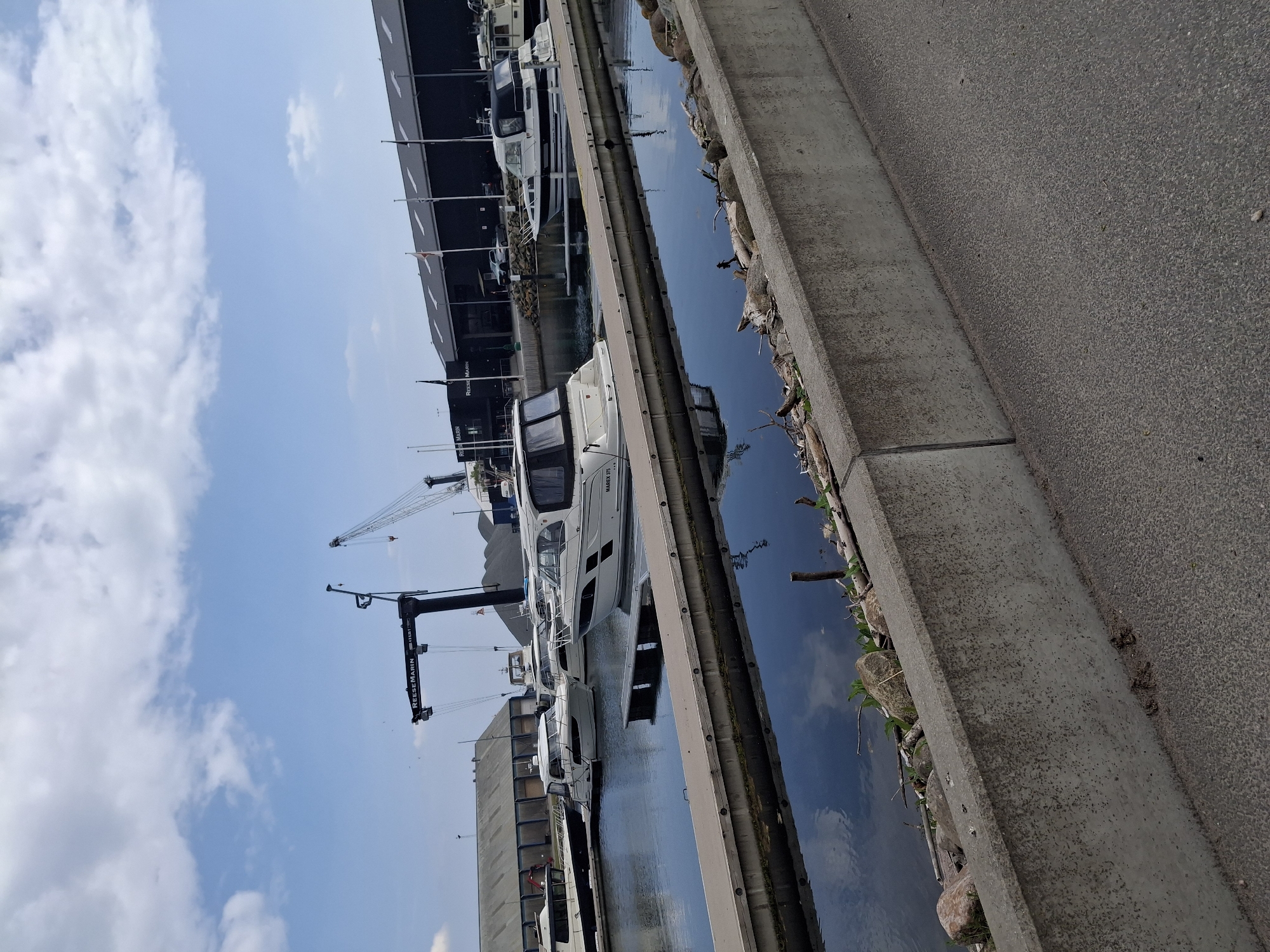 The image depicts a marina in Aabenraa, Denmark, with several yachts docked along a concrete pier. A large building, possibly a boat storage facility, is visible in the background. Cranes are present, suggesting boat maintenance and lifting capabilities. The sky is blue with scattered clouds. The image conveys a sense of activity related to boating and marine services.