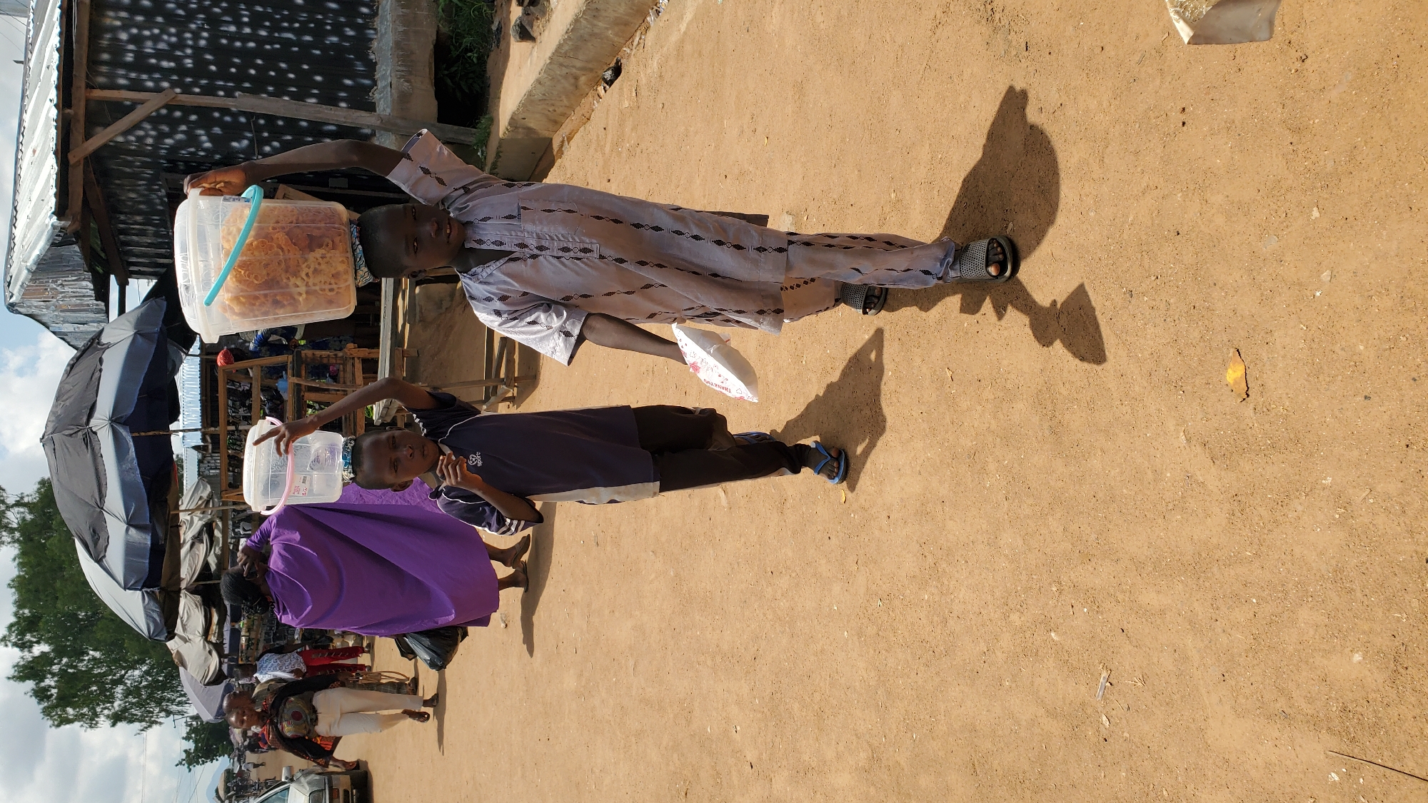 In Jalingo, Nigeria, two young boys walk on a dusty street carrying large plastic buckets of food on their heads. The boy on the left, wearing a dark blue and white shirt and black pants, has a clear bucket on his head and appears to be eating something from it. The boy on the right, dressed in a grey patterned tunic and pants, carries a bucket filled with what looks like puffed snacks or cereal. They are walking past market stalls, indicated by umbrellas and structures in the background. Other people and vehicles are visible in the distance, suggesting a bustling marketplace. The ground is composed of reddish-brown dirt, and the sky is partly cloudy.
