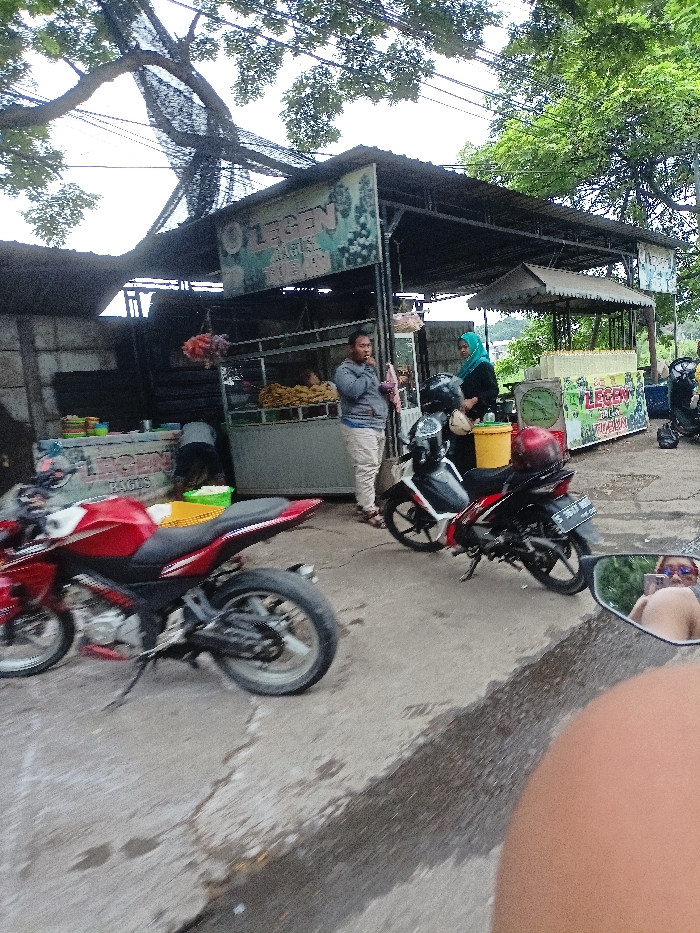 The image captures a vibrant roadside scene in Kecamatan Mojosari, Indonesia, centered around several outdoor food stalls operating under temporary shelters. Lush green trees and overhead utility lines are visible against the sky, suggesting a natural and built environment intersection.
In the foreground and midground, two motorcycles are prominent. On the left, a red sportbike is partially visible, parked on the concrete ground. To the right, a black and red scooter-style motorcycle, bearing the license plate "S 3073 MY," is parked near a stall, with a yellow bucket and a red helmet nearby. The photograph itself appears to be taken from a motorcycle, as indicated by a reflection of a person wearing sunglasses and revealing a bare shoulder in a rearview mirror at the bottom right.
Several individuals are present, engaging with the stalls. A man in a grey hooded top and light-colored pants stands near a stall displaying fried food items, looking towards the right. To his right, a woman wearing a green hijab and dark clothing stands beside the black scooter, facing the stalls. Another person is partially obscured as they bend down behind a counter on the left side of the scene. The overall activity suggests a typical daily market or street food interaction.
The stalls feature metal framework and corrugated roofs, common for outdoor vendors. A prominent sign above the central stall reads "LEGEN," with "ARIS" and "JUBAEDAH" visible below it, and "BAGUS" also discernable. Another smaller sign to the right also displays "LEGEN." The time of day appears to be daytime, with diffused lighting suggesting an overcast sky or late afternoon. The scene conveys a casual, bustling atmosphere typical of a local Indonesian street.