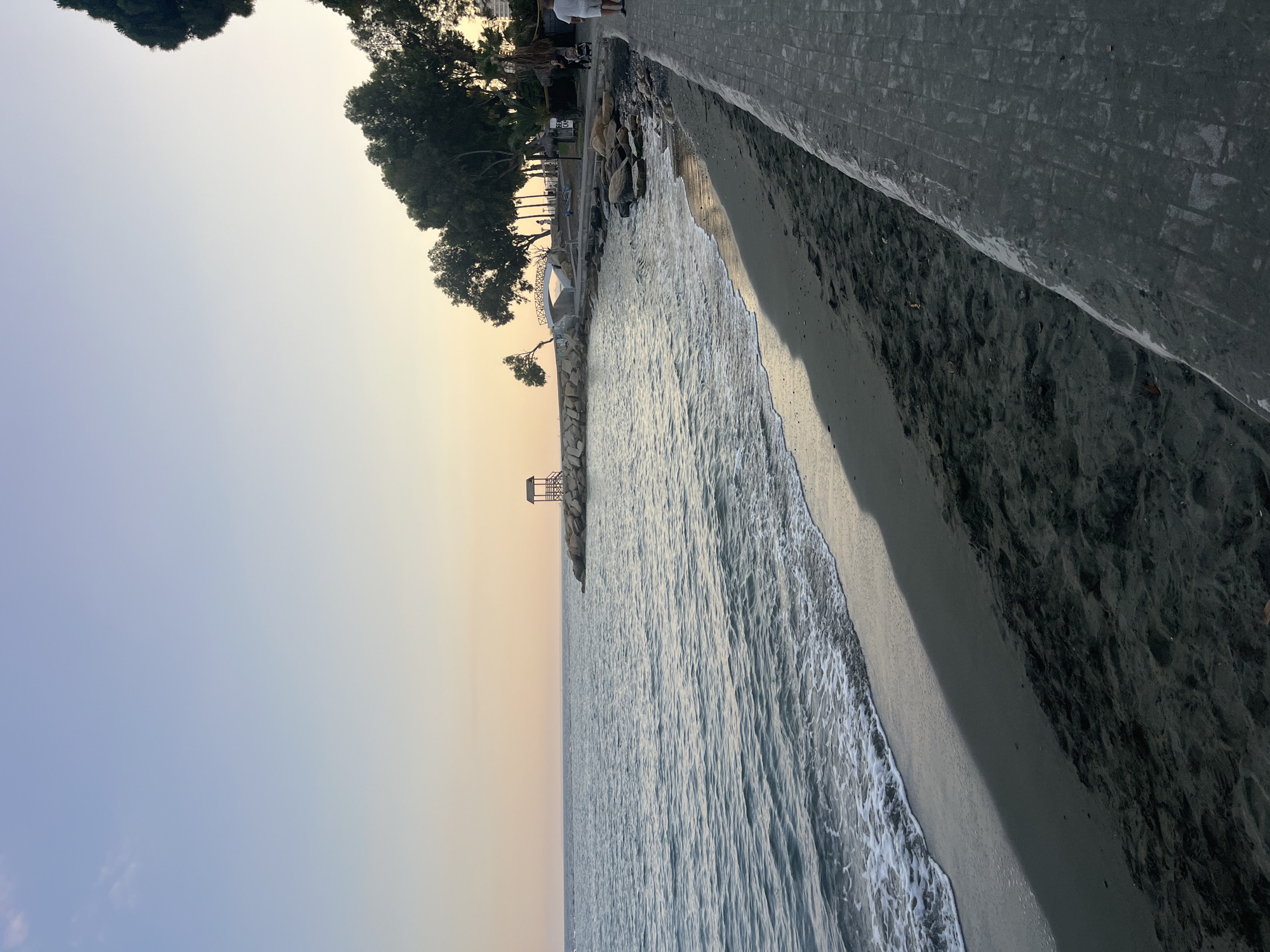 The image depicts a tranquil beachfront scene at dusk in the Agios Tychona Community, Cyprus. The sea, with gentle waves lapping at the dark sandy shore, occupies the left half of the frame. On the right, a paved walkway runs parallel to the beach, lined with rocks and vegetation. In the distance, a small wooden structure, resembling a lifeguard tower, stands on a rocky outcrop. Further along the coast, trees and a white arch bridge are visible, suggesting a developed area. The sky transitions from a pale blue at the top to a soft orange hue near the horizon, indicating sunset. A few individuals are present on the walkway; one person, wearing a white shirt and shorts, walks away from the camera, while another pushes a stroller. The overall atmosphere is peaceful and serene.