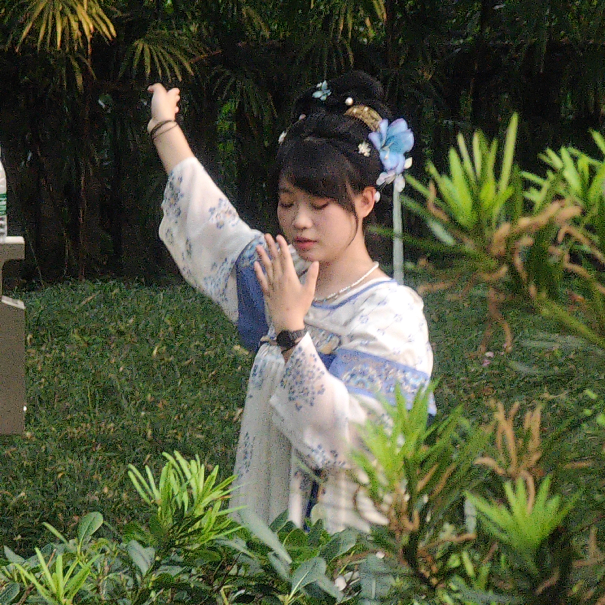 A young woman stands in a garden in Yuzhong District, China. She wears a traditional Hanfu dress with white and blue floral patterns, and her hair is styled in a traditional updo adorned with flowers. The woman is positioned slightly off-center, with green foliage framing her on both sides. She is raising one hand in a graceful gesture, while her other hand is closer to her body. She wears a watch on her left wrist and a pearl necklace. The background consists of various green plants and trees.