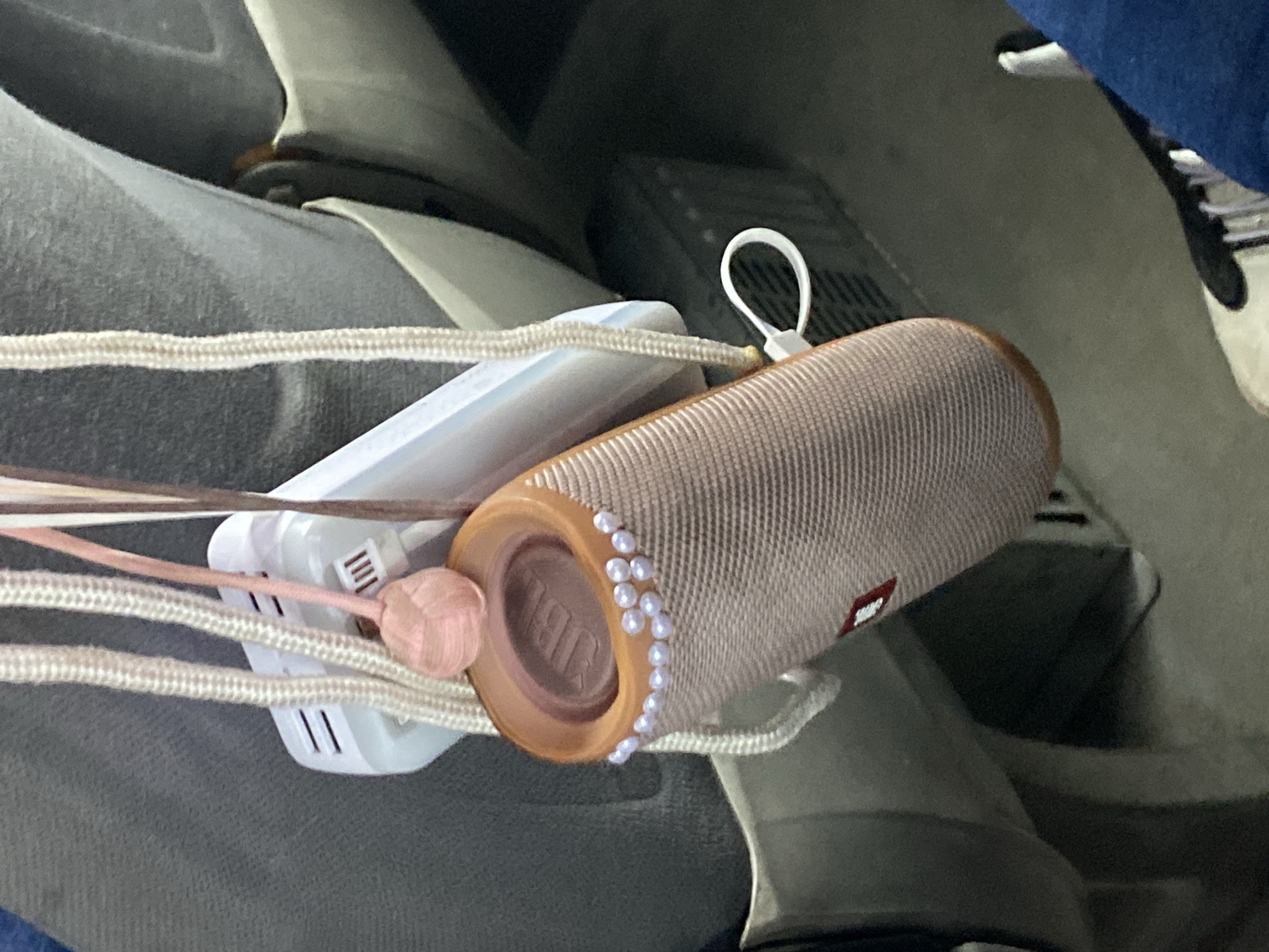 A close-up, high-angle shot shows a JBL Flip speaker, beige and adorned with a string of white pearls, lying inside a gray tote bag along with a white smartphone or charger. The bag has rope handles. The background shows parts of shoes and what appear to be car seats or furniture, suggesting this might be inside a vehicle or house. There is no indication that this image was taken in Krylovo, Russia, or even Russia.