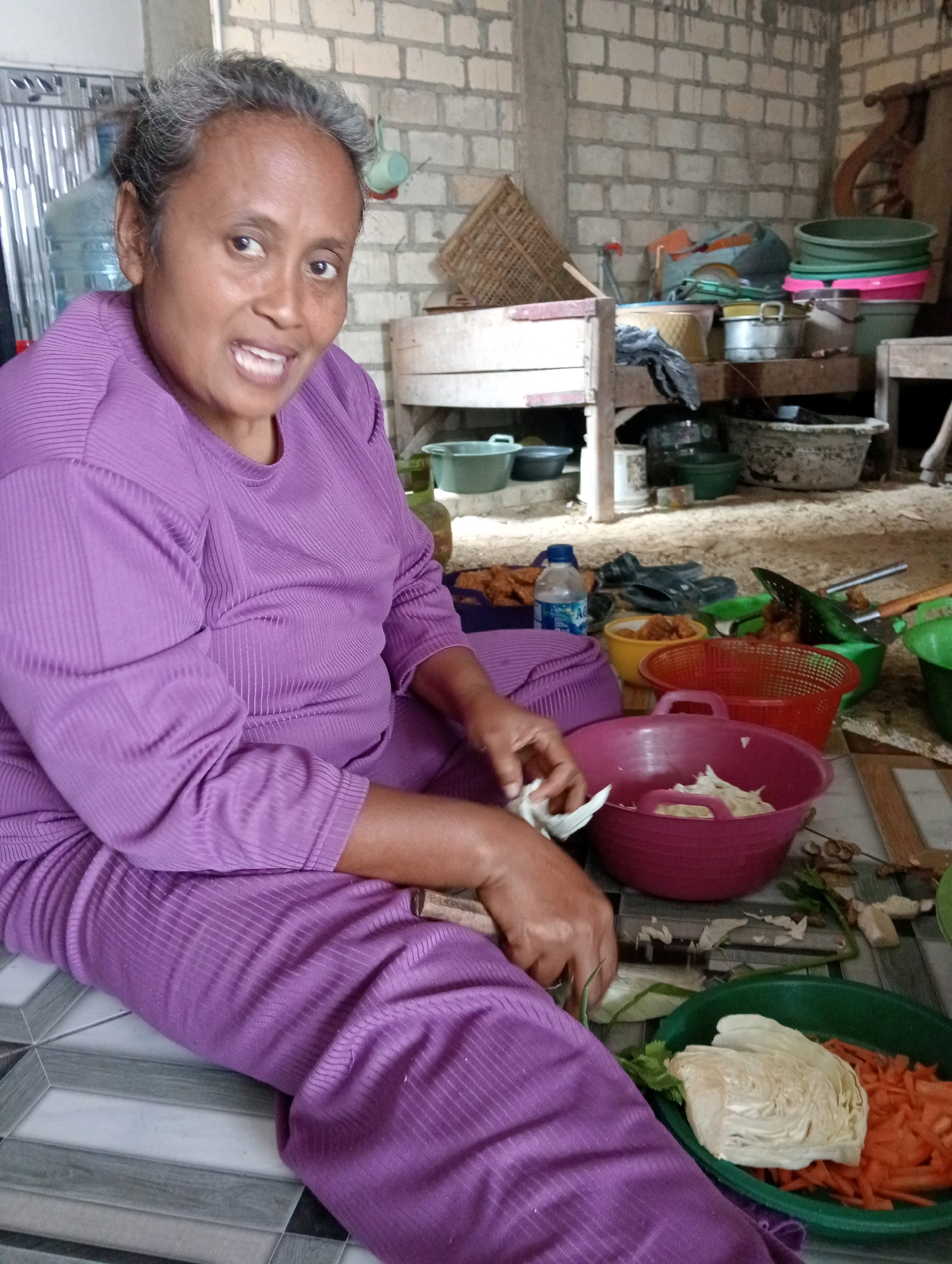 In Soko, Indonesia, a woman with graying hair and medium-brown skin, wearing a ribbed lavender-colored long-sleeved shirt and pants, sits on a tiled floor. She is facing the camera, with her mouth slightly open and her eyes looking forward. Her right hand is in the process of chopping or preparing white cabbage, holding a piece of it and a knife, over a pink plastic basin. In front of her, to her right, is a green plastic basin containing chopped carrots and a piece of cabbage. The background depicts a room with brick walls, wooden shelving filled with various containers, buckets, and bags, suggesting a functional, possibly outdoor or semi-enclosed, kitchen or storage area. The floor is covered with square tiles in a grey and white pattern.