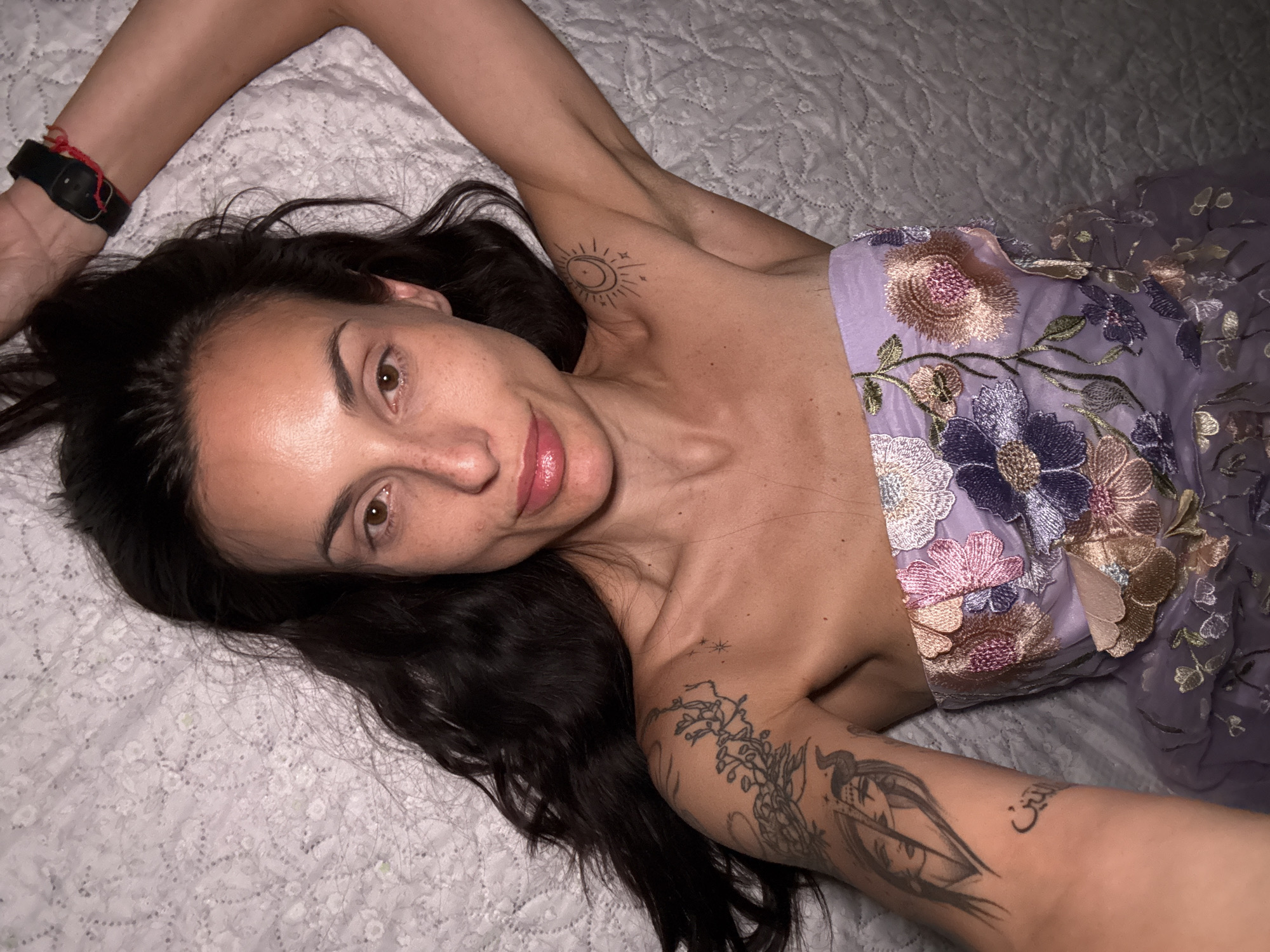 A woman lies on a white textured surface. She is wearing a purple strapless dress with floral embroidery. Her dark hair is spread around her head on the white surface. She has tattoos on both arms and on her chest. She is facing the camera with her arms raised on either side of her head. The location is not discernible from the image. There is no indication that this image was taken in Pleven, Bulgaria.