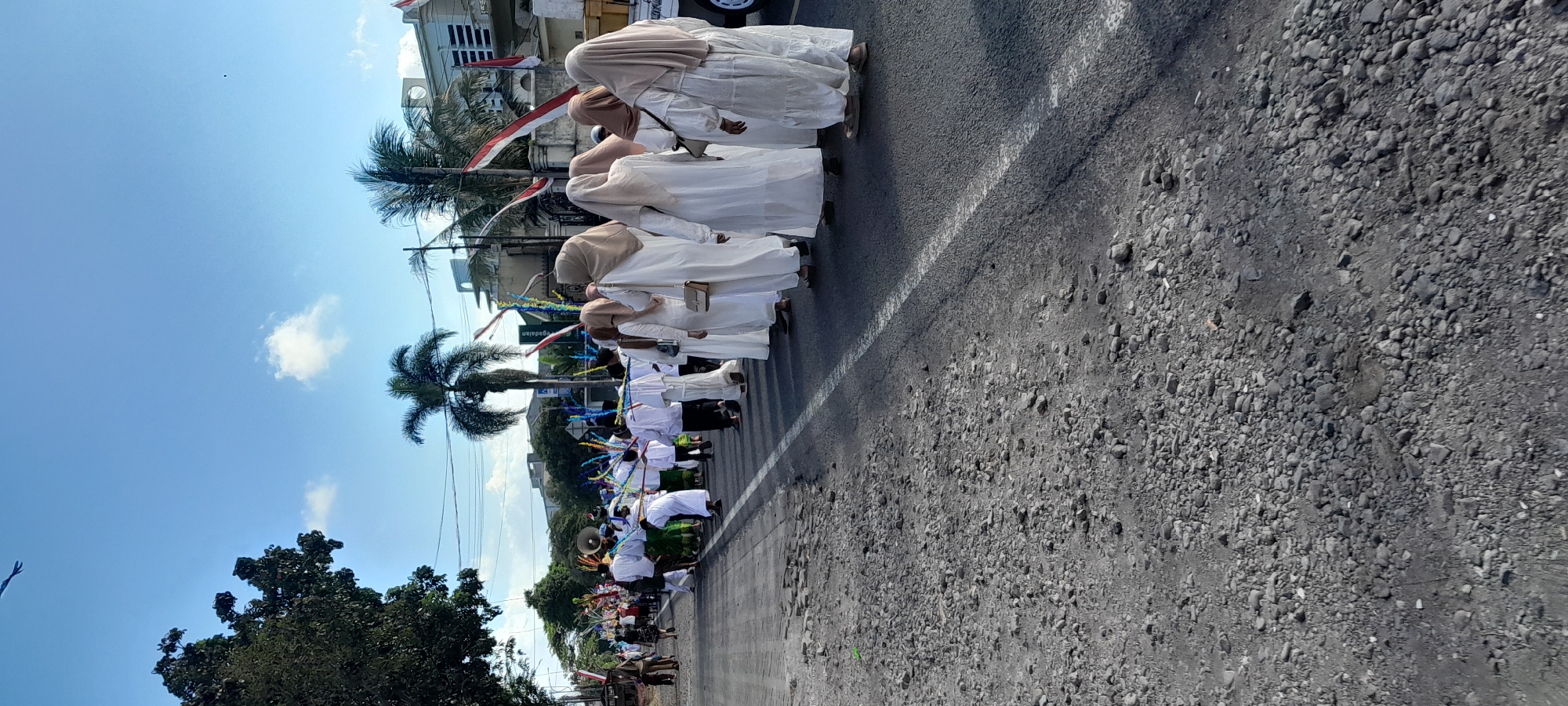 In Rogojampi, Indonesia, a procession moves along a paved road. The group consists of individuals dressed in white robes and headscarves, some carrying long, decorated poles. In the background, palm trees and buildings are visible under a clear blue sky with scattered clouds. The road is bordered by a gravelly shoulder and appears to be a public street within a city.