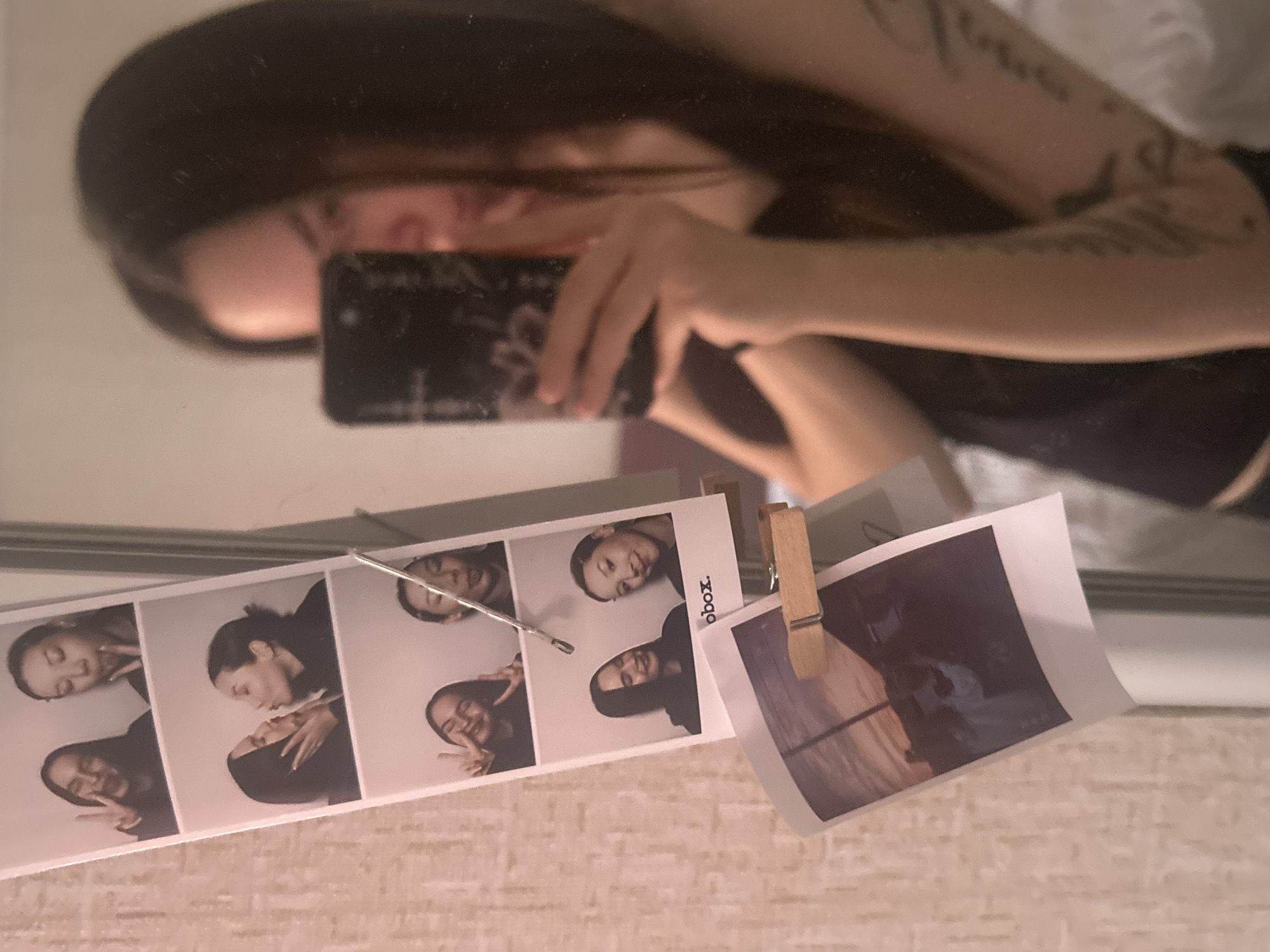 A person with long, dark hair and tattoos on their arm takes a mirror selfie using a black smartphone. The mirror reflects their face, arm, and part of their body, which is dressed in a dark-colored top. The mirror is fixed to a surface, probably a wall. On the surface, there is a strip of four photo booth pictures of a woman with dark hair, likely the same person. The photos depict her making different facial expressions and poses. Beside the photo strip, a photograph is attached to the mirror's edge with a small wooden clothespin. The photograph's subject is difficult to discern. The background behind the mirror appears to be a wall or another surface with a light-colored, textured pattern. The location, Chaikovsky, Russia is not discernible from the image's visual data.
