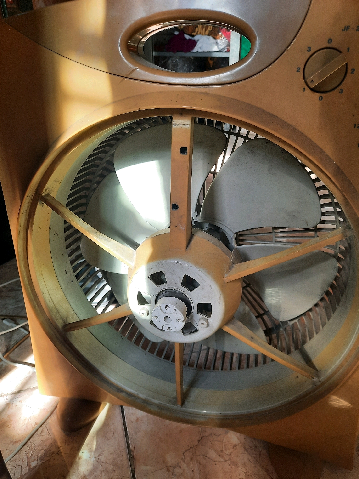 This is a close-up, medium shot of the front grille and fan blades of an orange electric fan, positioned indoors. The fan is dusty, and the blades are a light gray color. The fan has a rotary dial on its upper right side with settings numbered 0, 1, 2, and 3. A light source, likely sunlight, casts strong shadows and highlights across the fan and the floor, which appears to be tiled. The background shows a glimpse of items inside what looks like a closet or shelf, including clothing. The image does not provide any specific information about its location within East Jakarta, Indonesia, or any people present.