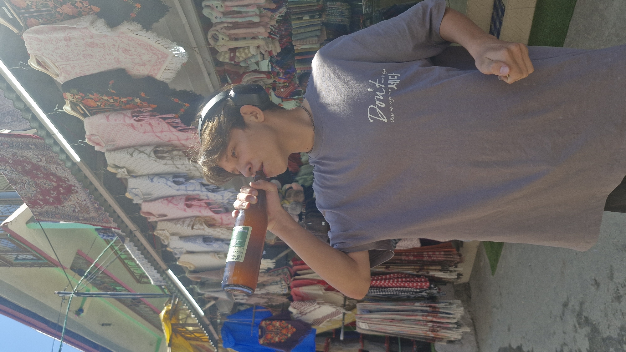 In Vashisht, India, a young person wearing a grey t-shirt and headphones drinks from a glass bottle. The t-shirt has white text that reads "Don't make the days count, make the days count," followed by Korean characters. The person is standing on a paved area in front of a market stall displaying colorful textiles, scarves, and blankets. The market stall is set up outdoors, under a covered structure with bright lighting. Behind the stall, a building with balconies and colorful paint is visible. The scene is brightly lit by sunlight, indicating it is daytime.