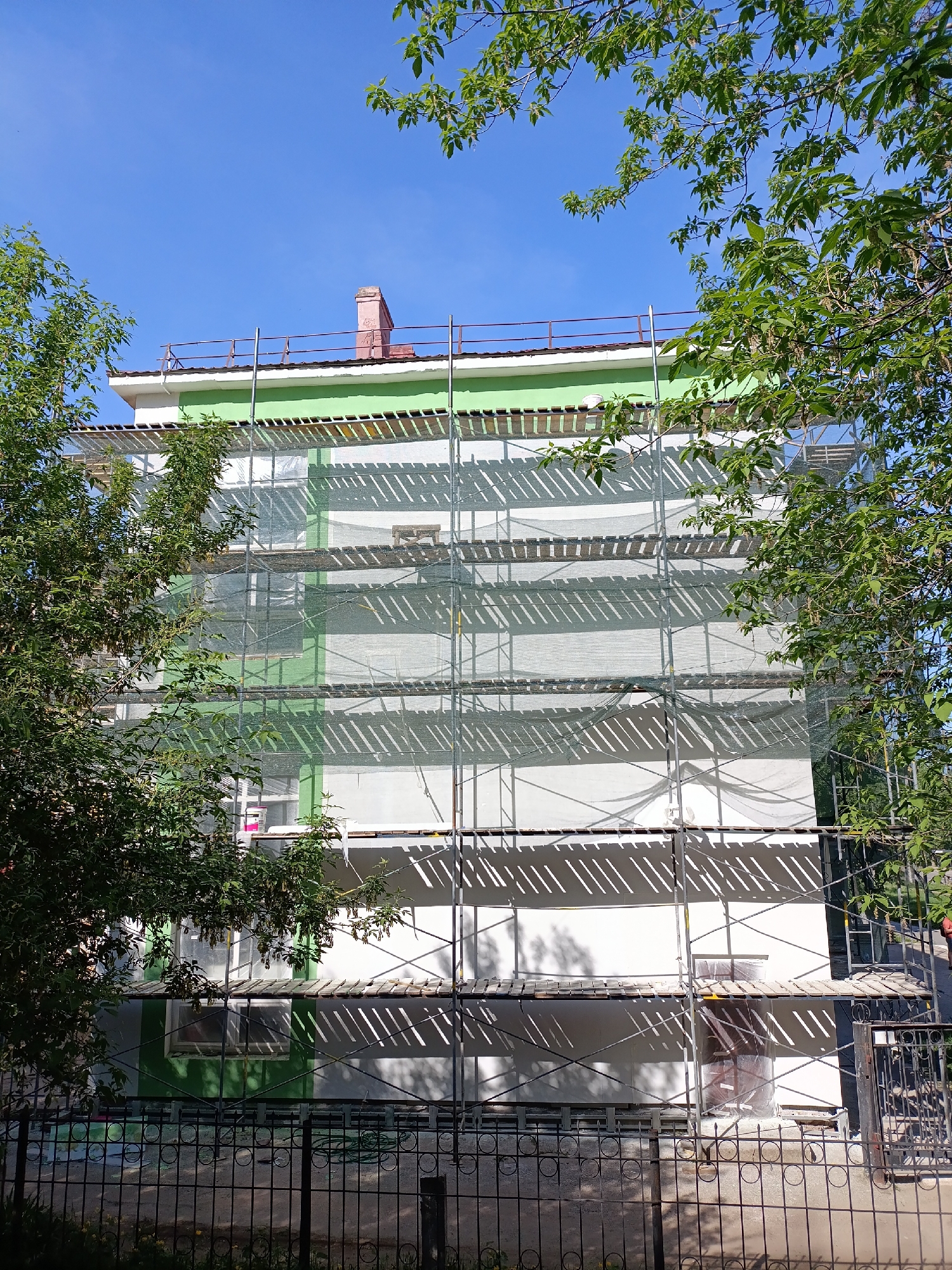 The image shows a multi-story building in Chaikovsky, Russia, undergoing renovation. Scaffolding covers the building's facade, partially obscured by netting. The building is painted in shades of green and white. A red brick chimney is visible on the roof. The foreground includes a black iron fence and foliage from trees framing the scene. The sky is clear and blue.