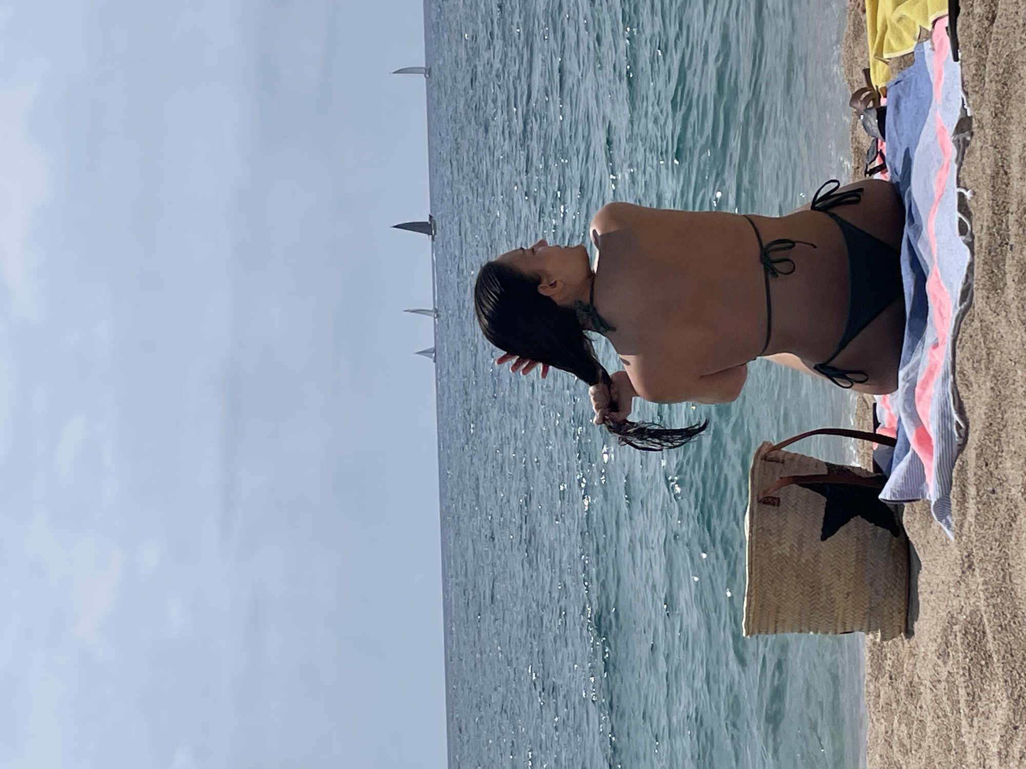 The image depicts a woman sitting on a sandy beach in Canet de Mar, Spain, facing the ocean. She is wearing a dark green bikini and is in the process of brushing her wet, dark hair with her hands. In the background, several sailboats are visible on the shimmering blue water under a hazy sky. A woven straw bag with a black star detail and a striped towel are placed on the sand next to her. The scene captures a relaxed moment by the sea.