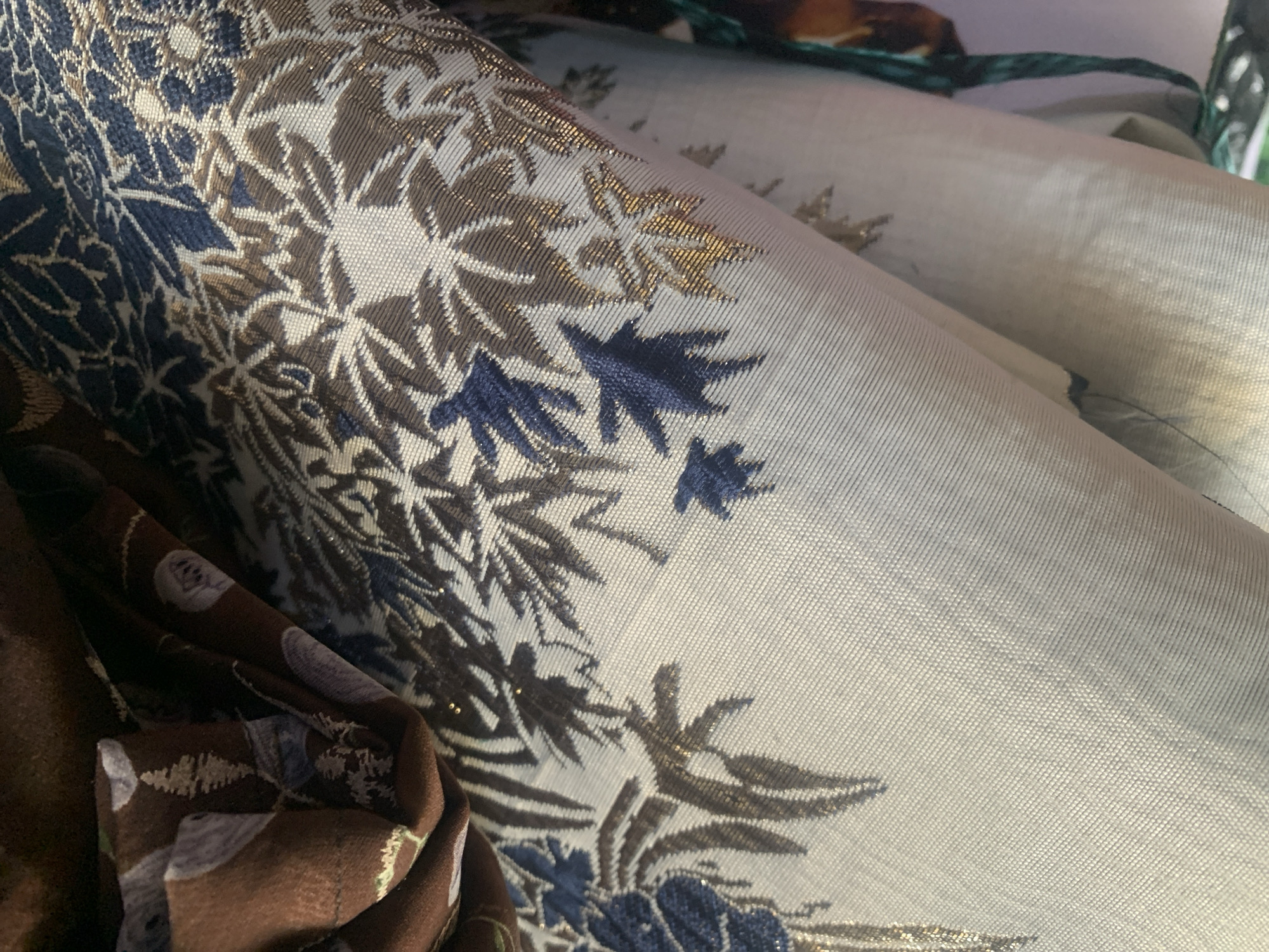 The image shows a close-up view of patterned fabric, likely a textile or garment. The fabric features a design with floral and leaf motifs in shades of blue, brown, and gold, against a light-colored background. The texture of the fabric appears to be woven, with some areas possibly having a metallic sheen due to the gold threads. The fabric is folded and draped, creating shadows and highlights that emphasize its texture and pattern. The background is out of focus, suggesting the image was taken in a setting where such materials are displayed or stored. There is no discernible urban or environmental context to place this image within Jauro Yinu, Nigeria. The image solely focuses on the detail and texture of the fabric.