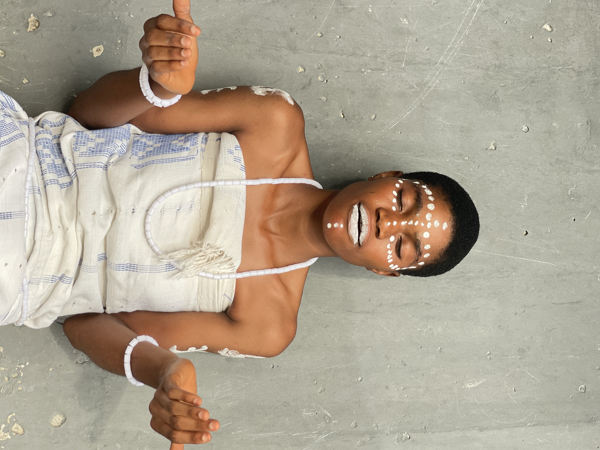 A person lies supine on a gray surface. They wear a white garment with blue stripes, a white beaded necklace, and white bracelets. White paint is applied in dots to their face and in patches on their shoulders. The location is likely Badagry, Nigeria, based on the subject's apparent cultural attire. The person's eyes are closed and their lips are painted white. The hands are raised, with fingers gesturing. The surface beneath the person shows signs of wear, with marks and small debris visible.