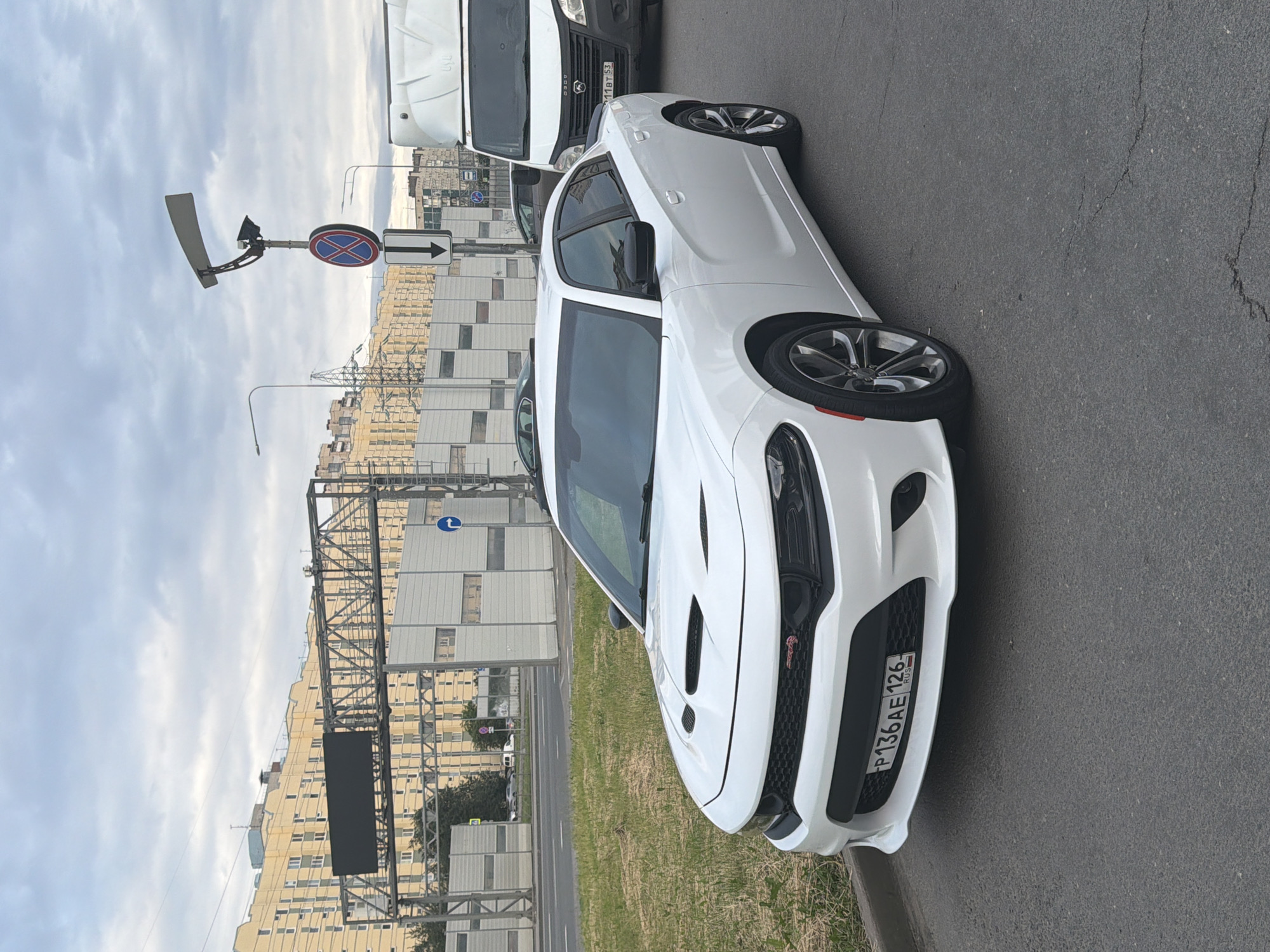 A white Dodge Charger is parked on the right side of an asphalt road in an urban setting. The car is facing slightly to the left, with its front prominently displayed. The license plate on the car reads "P136AE 126". To the right of the Charger, a white commercial van is parked. In the background, there are buildings, a highway overpass with an electronic display, and a traffic light with a circular "no stopping" sign. The sky is overcast. The ground is paved and cracked in places.