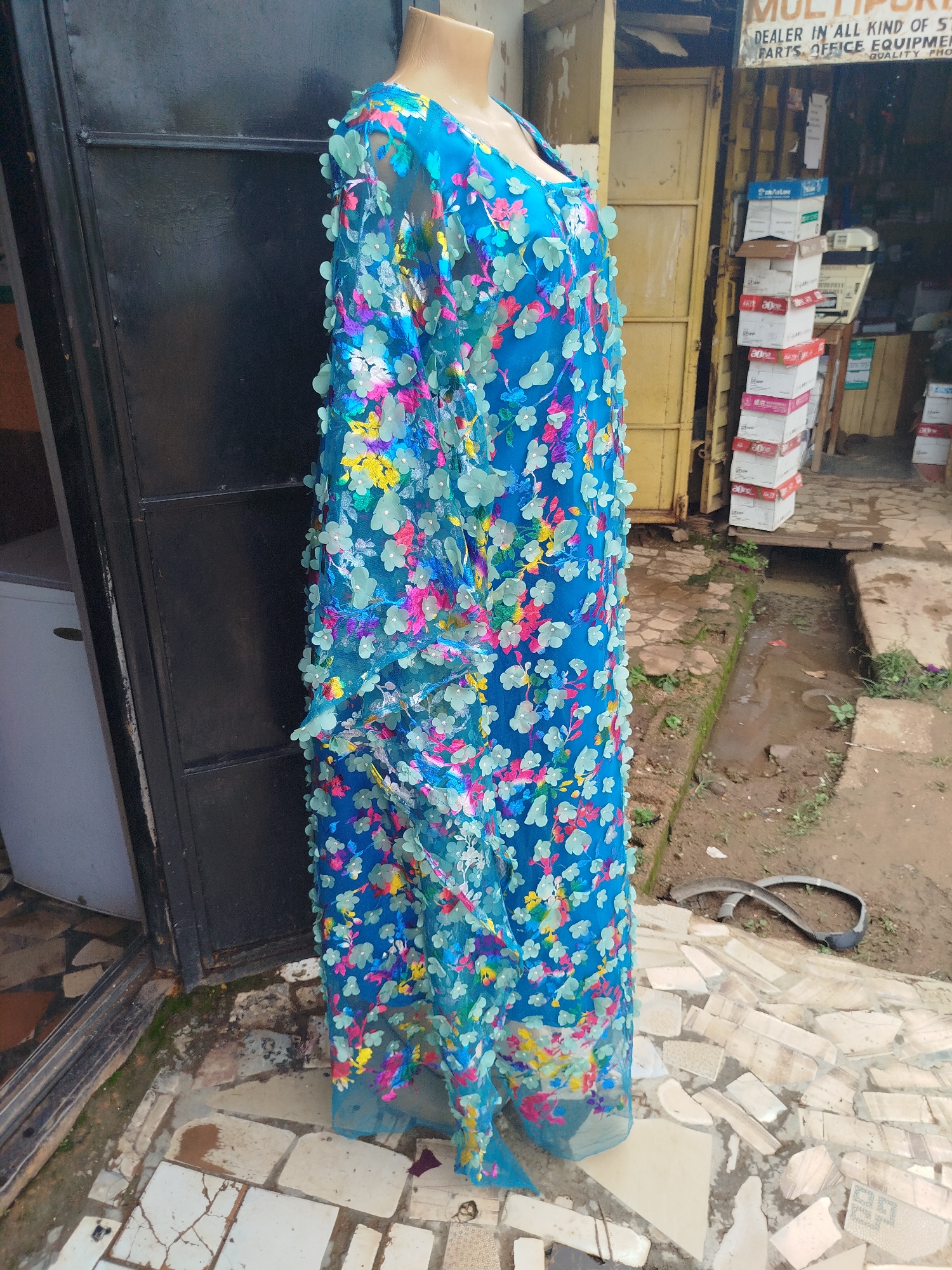 A mannequin displays a long, blue, sheer dress adorned with colorful floral appliques in a street setting in Rafin Par, Nigeria. The dress has a V-neck and appears to be made of lace or a similar delicate fabric, with light blue, pink, yellow, and green flowers scattered across it. The mannequin stands on a tiled walkway in front of a dark, corrugated metal door. To the right of the mannequin, a building with a yellow door and a sign that reads "MULTIPR..." is visible, with stacks of boxes and a white machine on display outside. A small, muddy ditch runs along the edge of the tiled walkway. The ground is a mosaic of cracked and uneven tiles.
